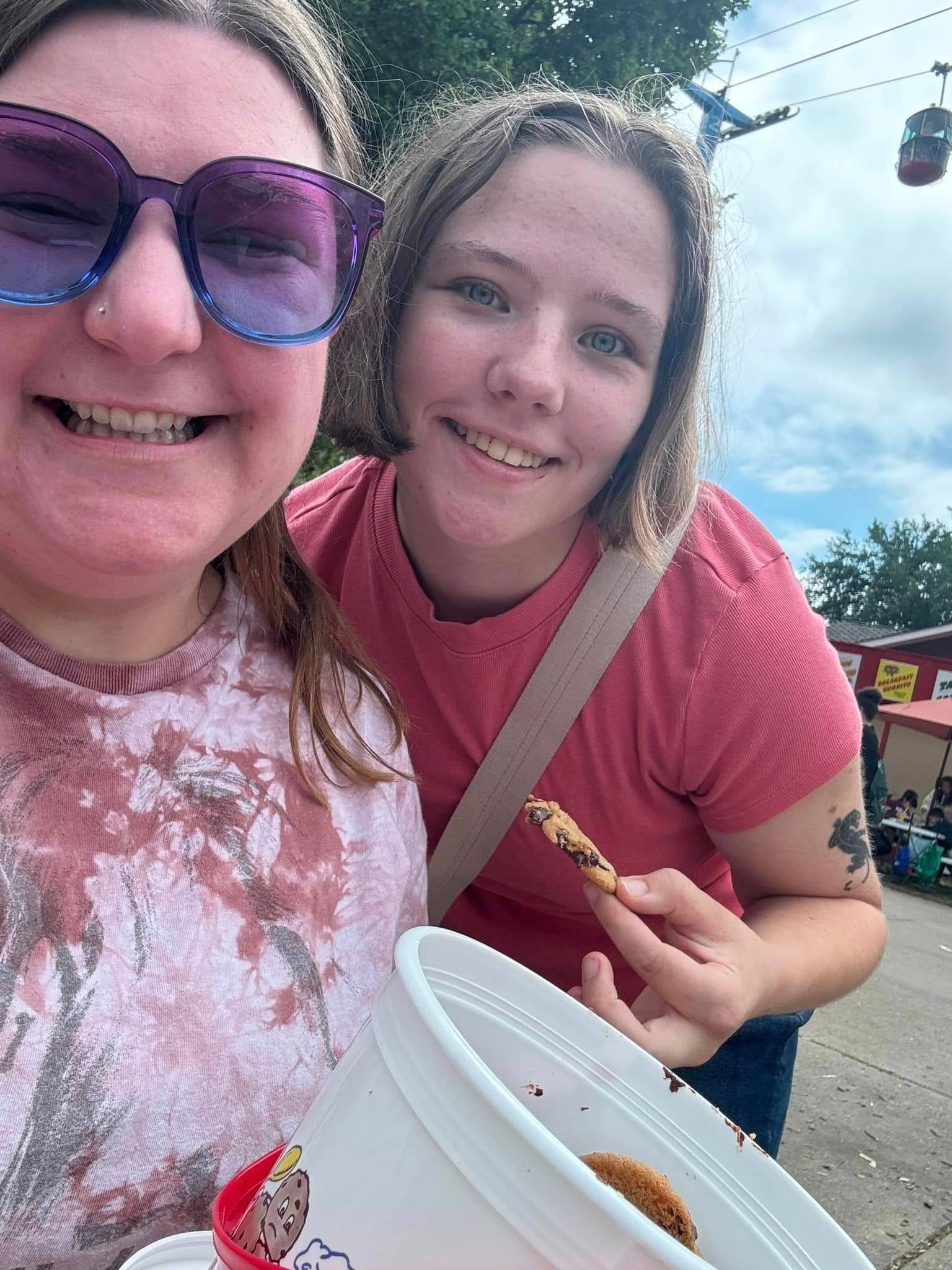 Two smiling people at a fair. One holds a snack, other wears sunglasses, and both smile at camera, blue sky background.