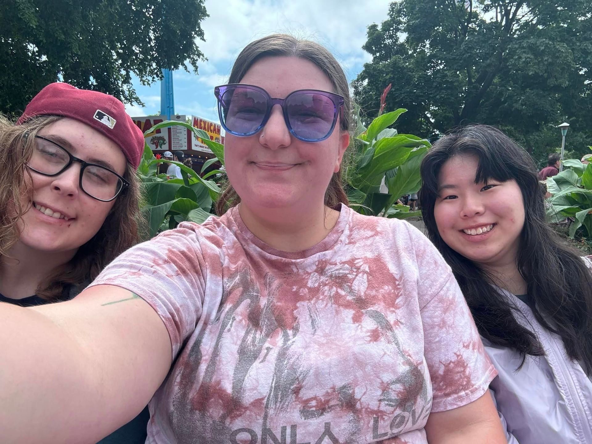 Three people smiling for a selfie outdoors. Center person wears sunglasses and tie-dye shirt. Trees and sky in background.