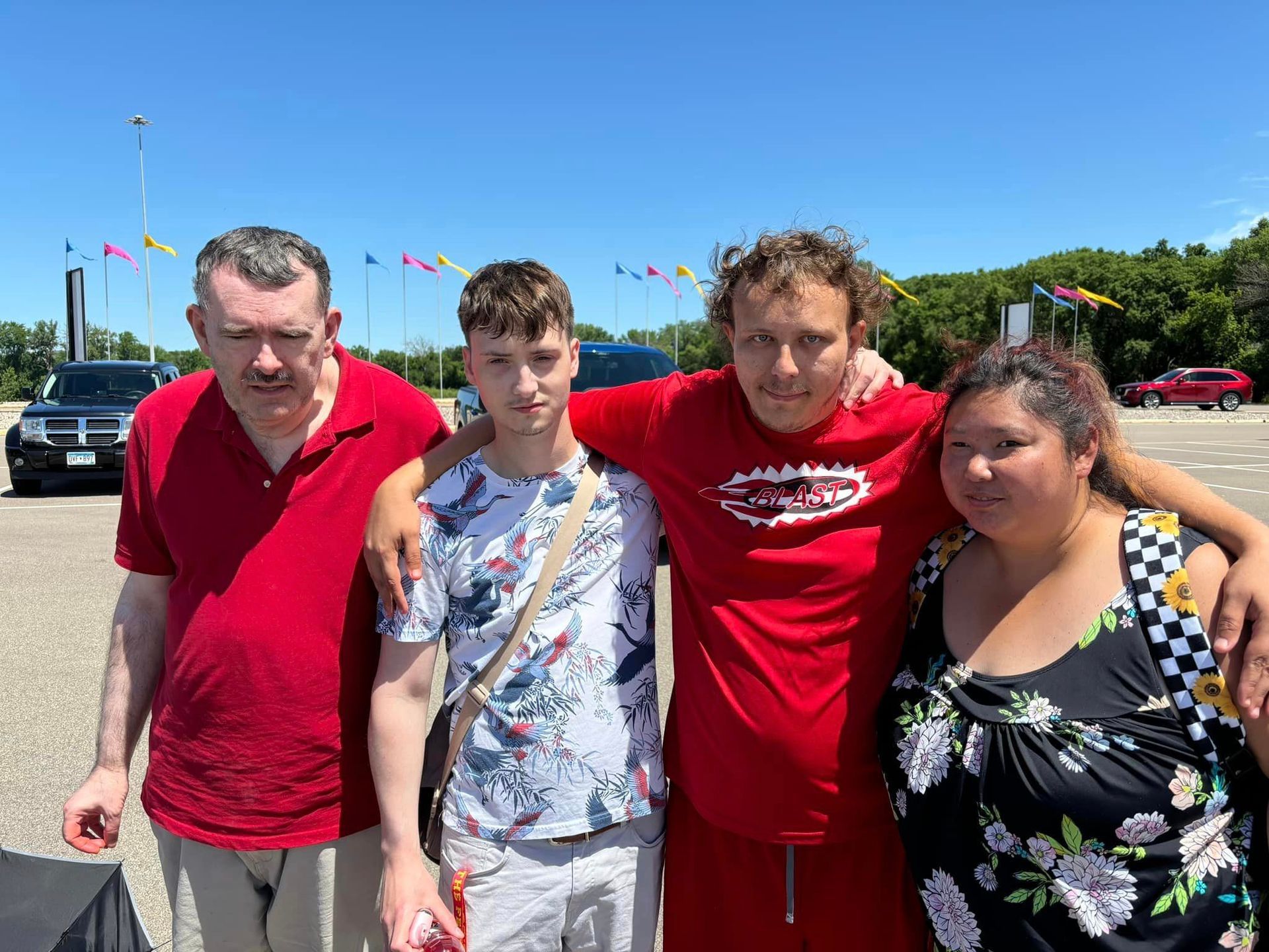 Four people standing outdoors. Two have arms around each other. Bright sunny day, blue sky.