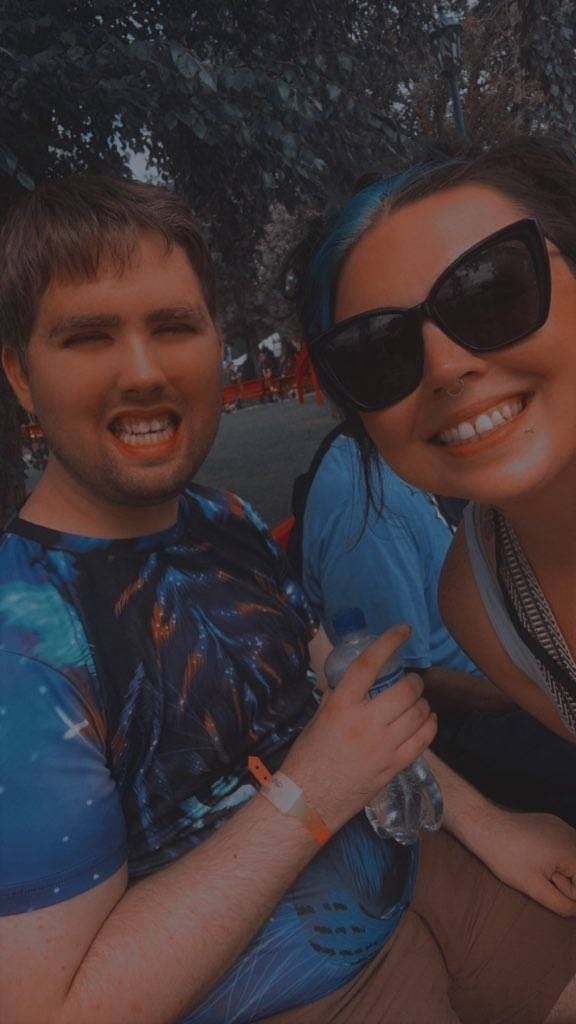 Two people smiling, one with blue hair and sunglasses, the other with a galaxy print shirt, outdoors.