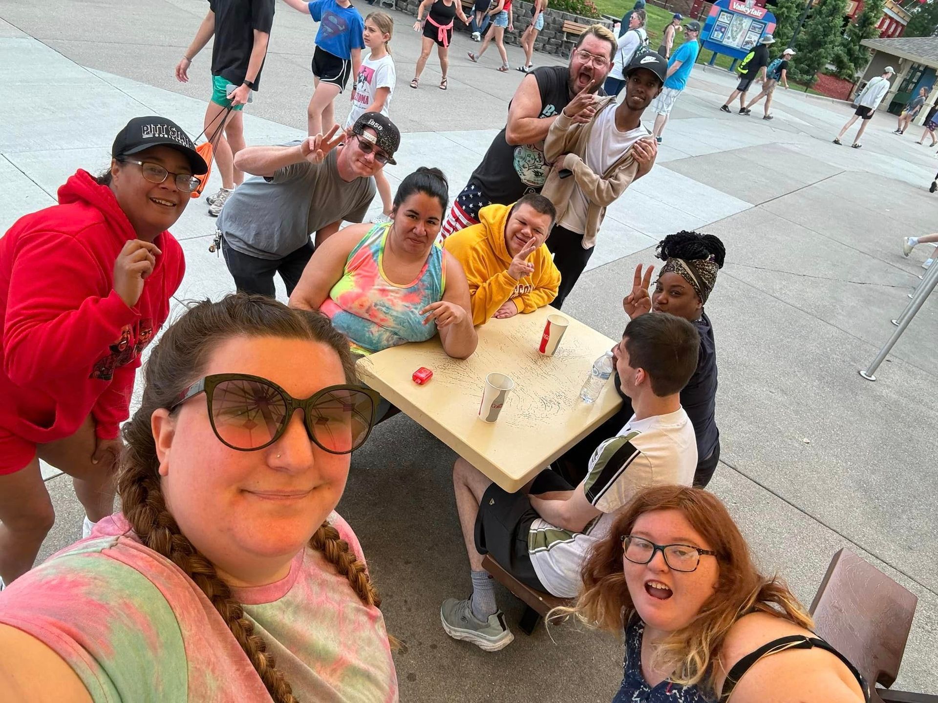 Group of people smiling, posing around a picnic table, outdoor setting. Some are making peace signs.