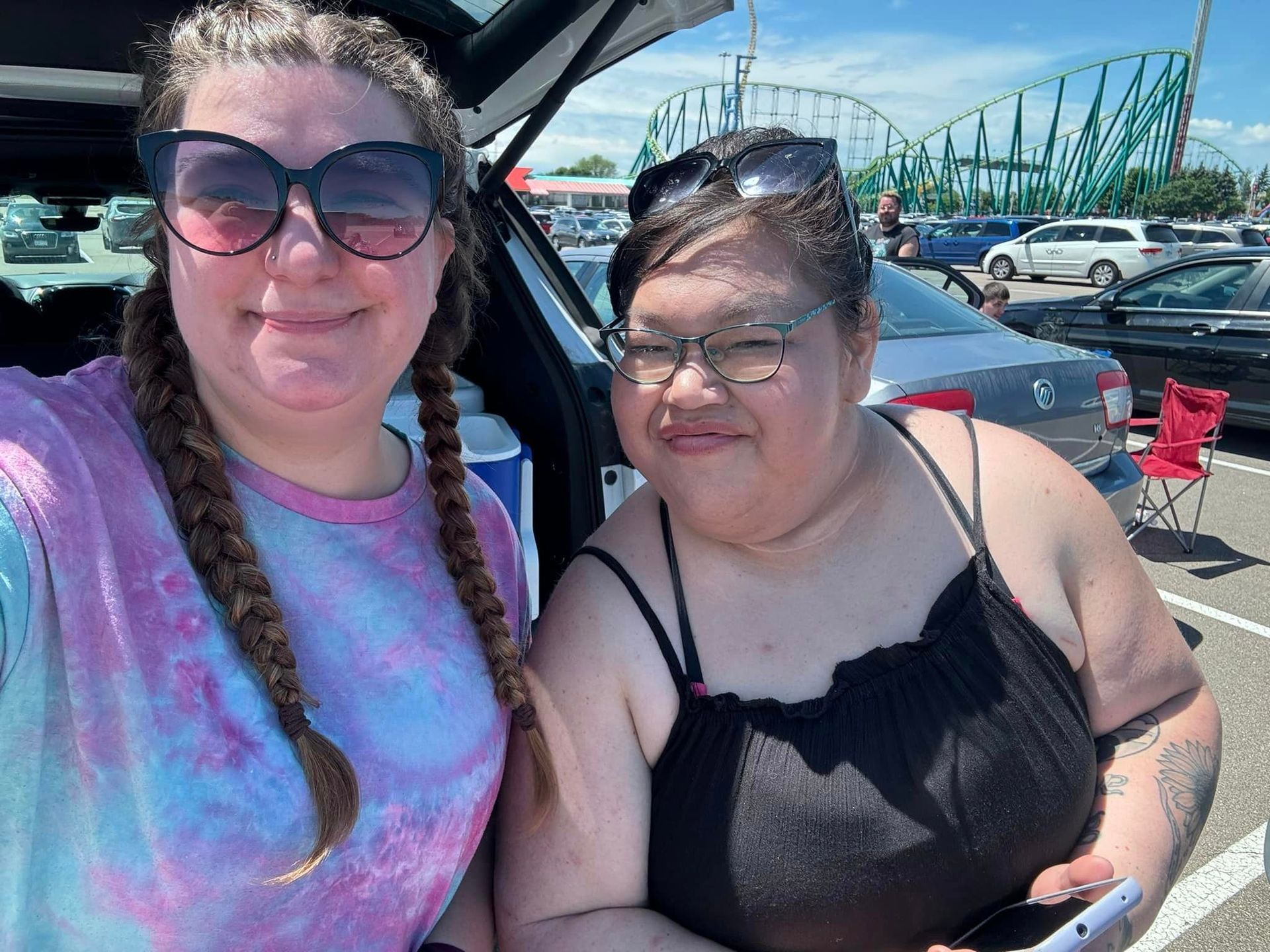 Two smiling people near a car, one with sunglasses and braids, the other wearing glasses and holding a phone