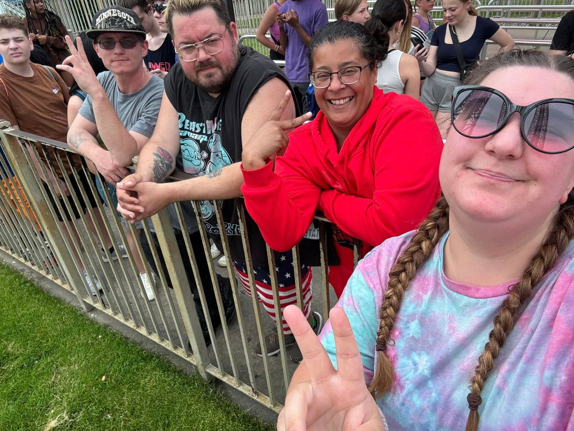 People smiling at the camera near a metal fence. One person wears tie-dye, others are behind the fence.
