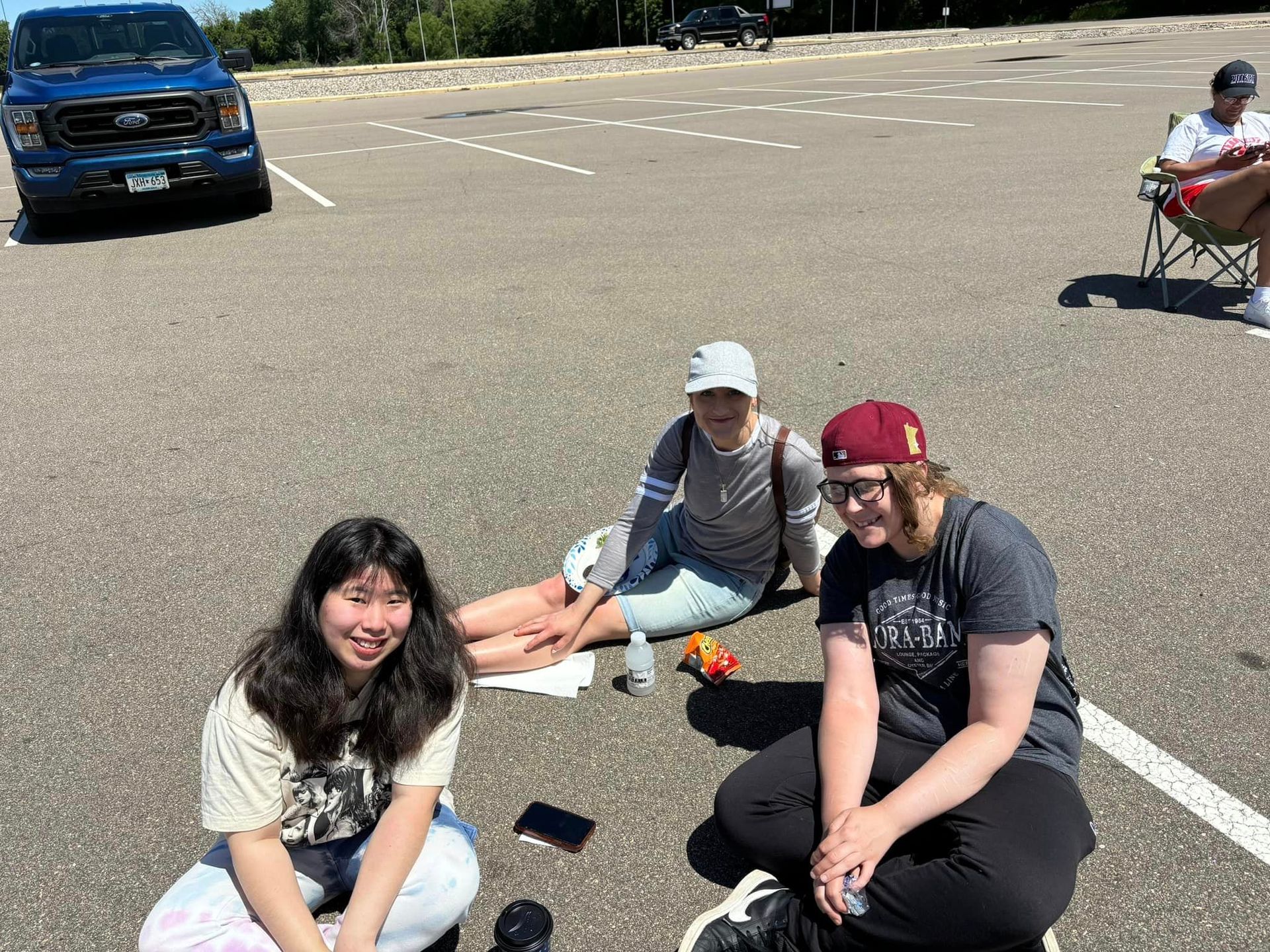 Three people sit on pavement, one looking at the camera and smiling. A blue truck and another person are visible.