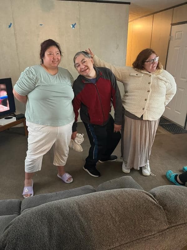 Three people posing playfully indoors; one leans head sideways, others smile, wearing casual clothes.