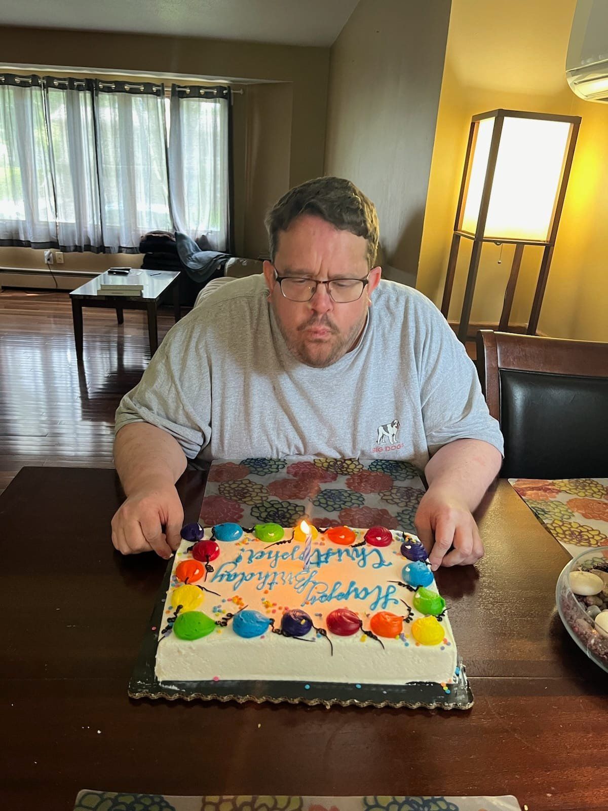 Man blowing out a birthday candle on a decorated cake at a table. Room setting with window and lamp.