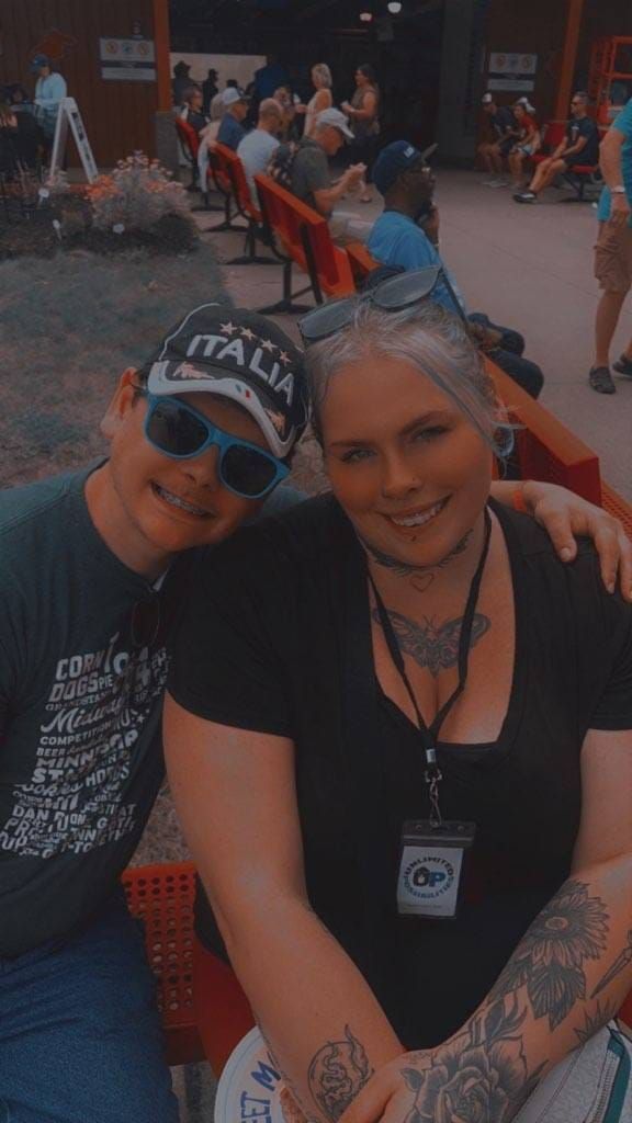 A woman and a man smile, posing for a photo outdoors. The woman has a lanyard and tattoos.