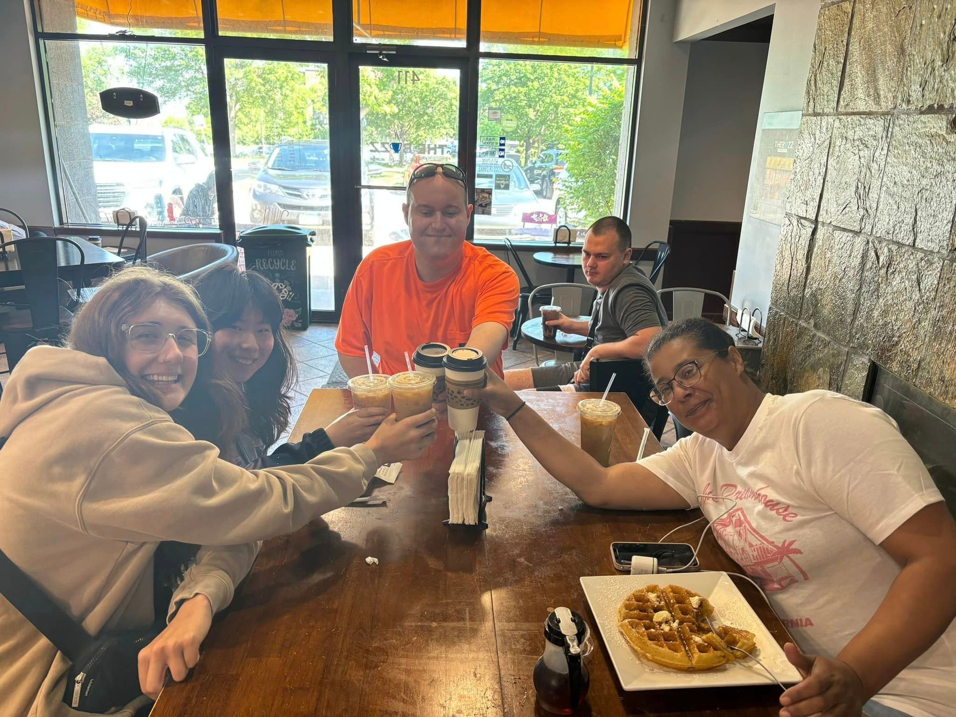People toasting treats at a cafe table.  Sunny window, waffle, and drinks are visible.