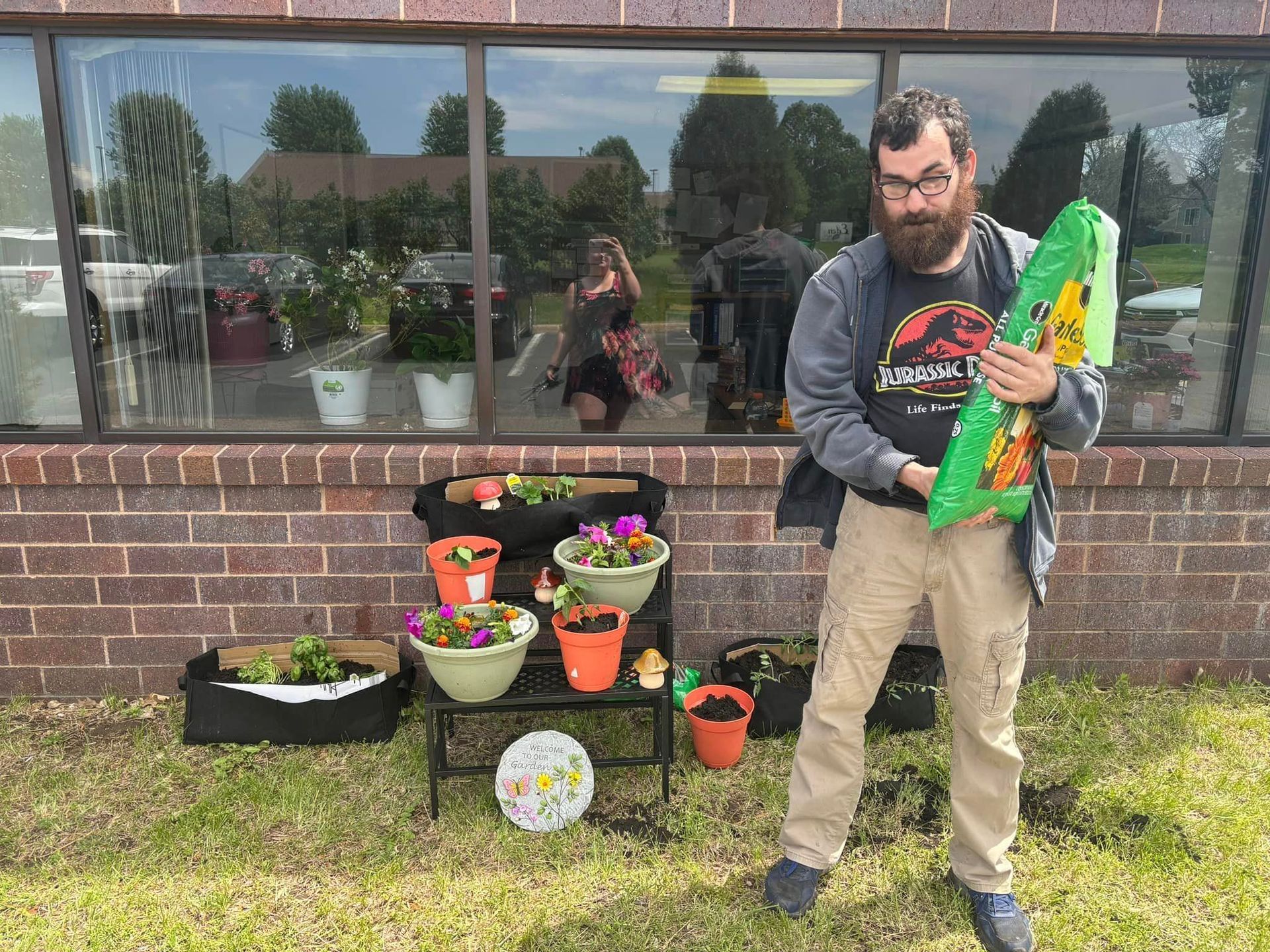 Man holding soil bag near potted plants in front of a brick building.
