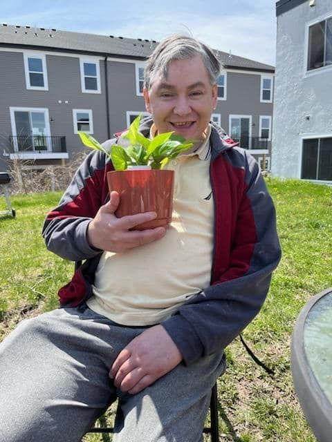 Man holding a potted plant, smiling in a backyard on a sunny day.