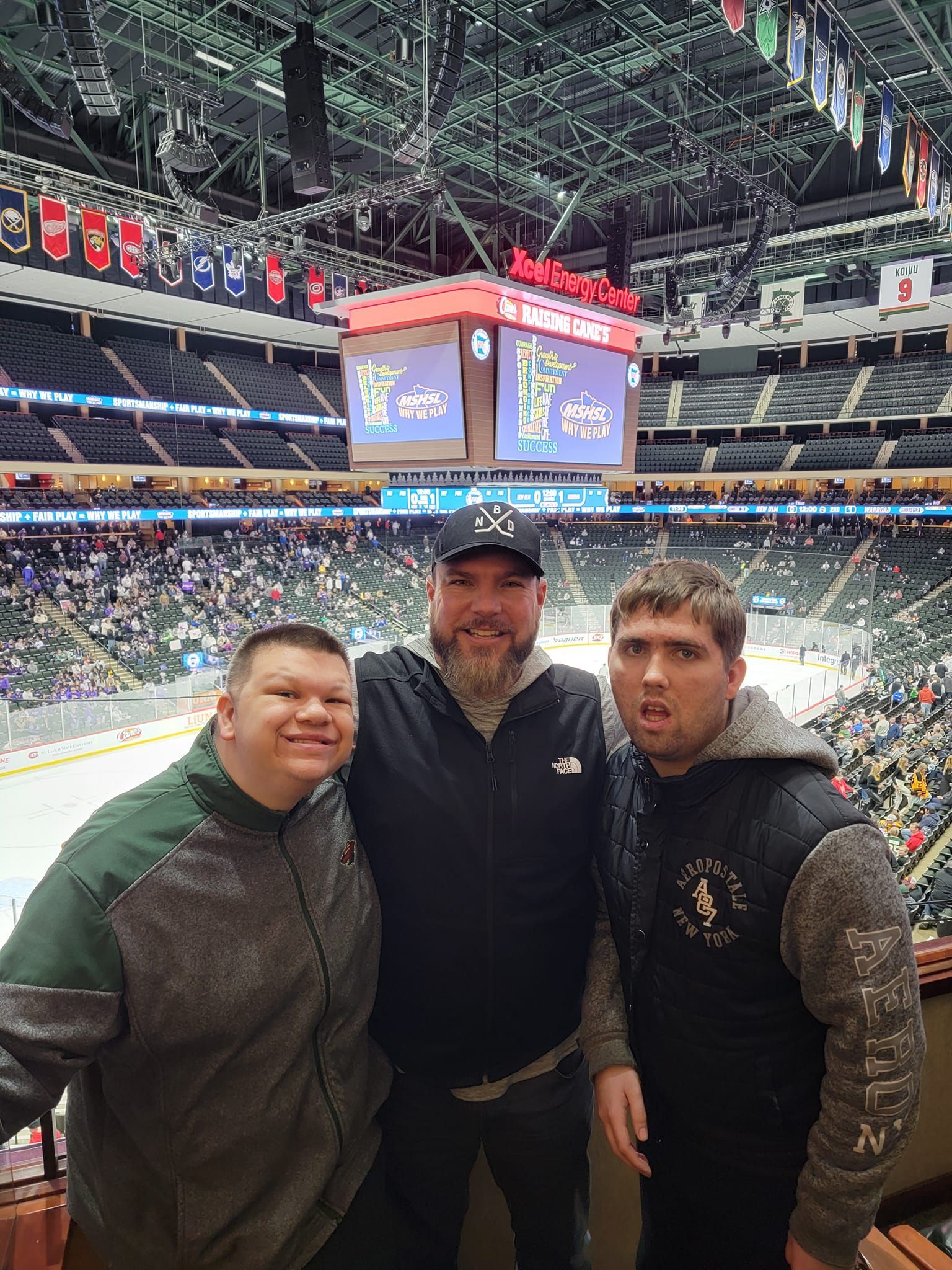 Three people smiling at an ice hockey game. They stand in front of a stadium full of fans, scoreboard visible.