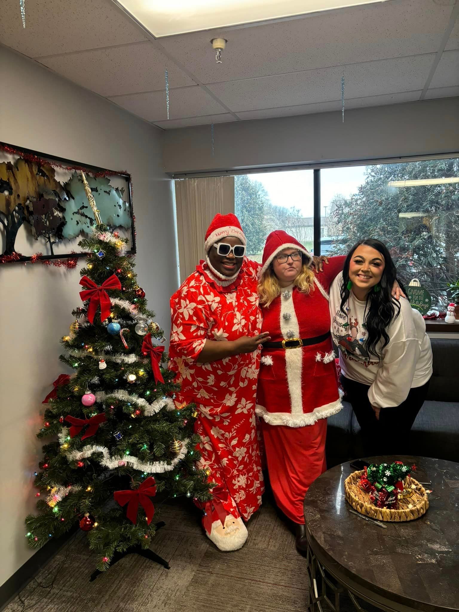 Three people pose by a Christmas tree in an office. Two wear Santa costumes and one a festive white top.
