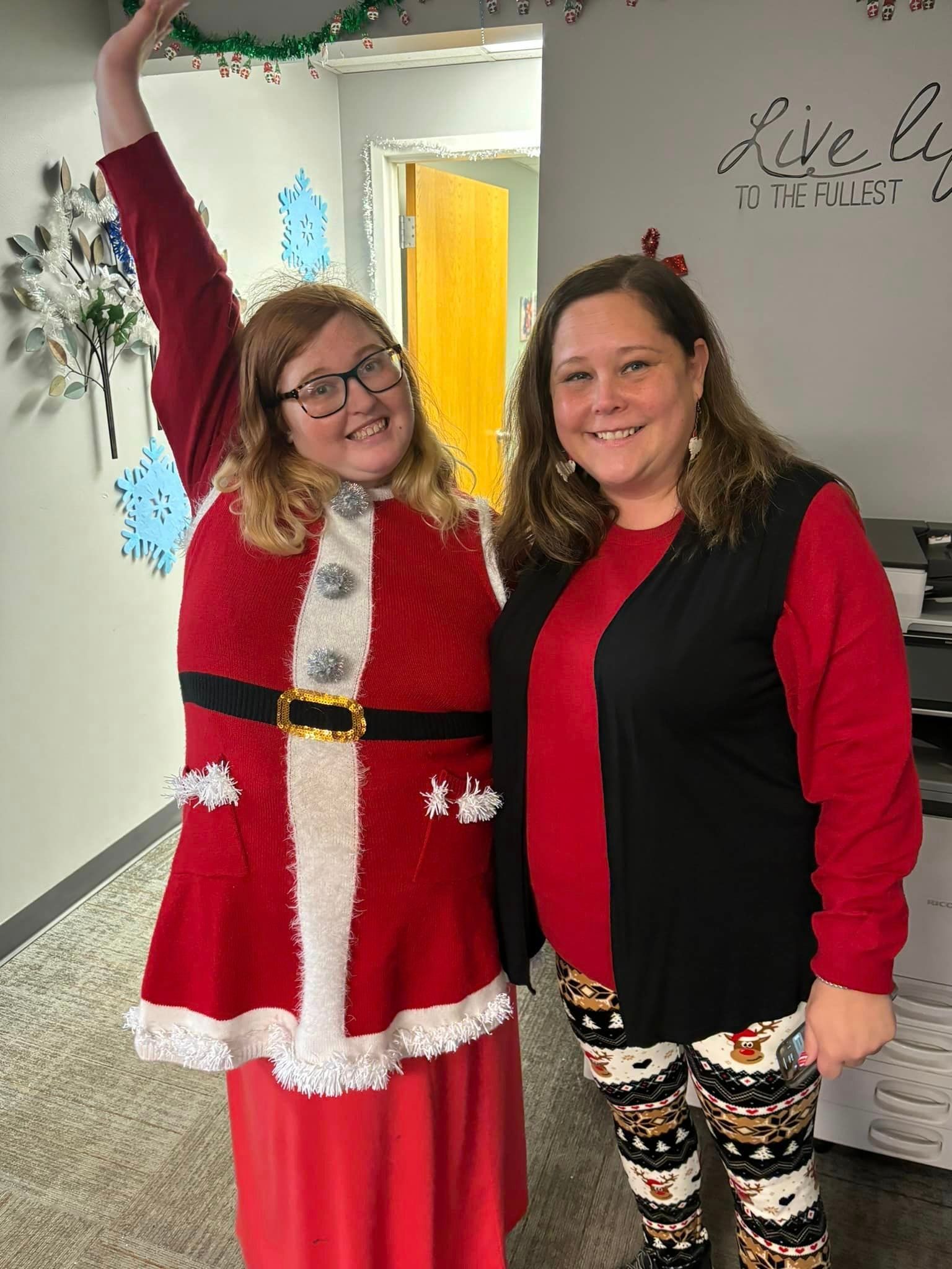 Two women in festive attire, one in a Santa suit, smiling, indoors.