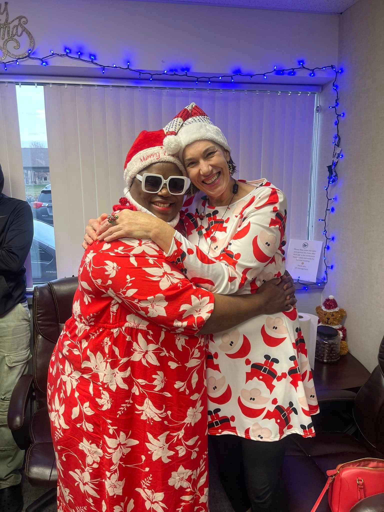 Two women in Santa hats and holiday outfits hug indoors, with blue lights and a window.
