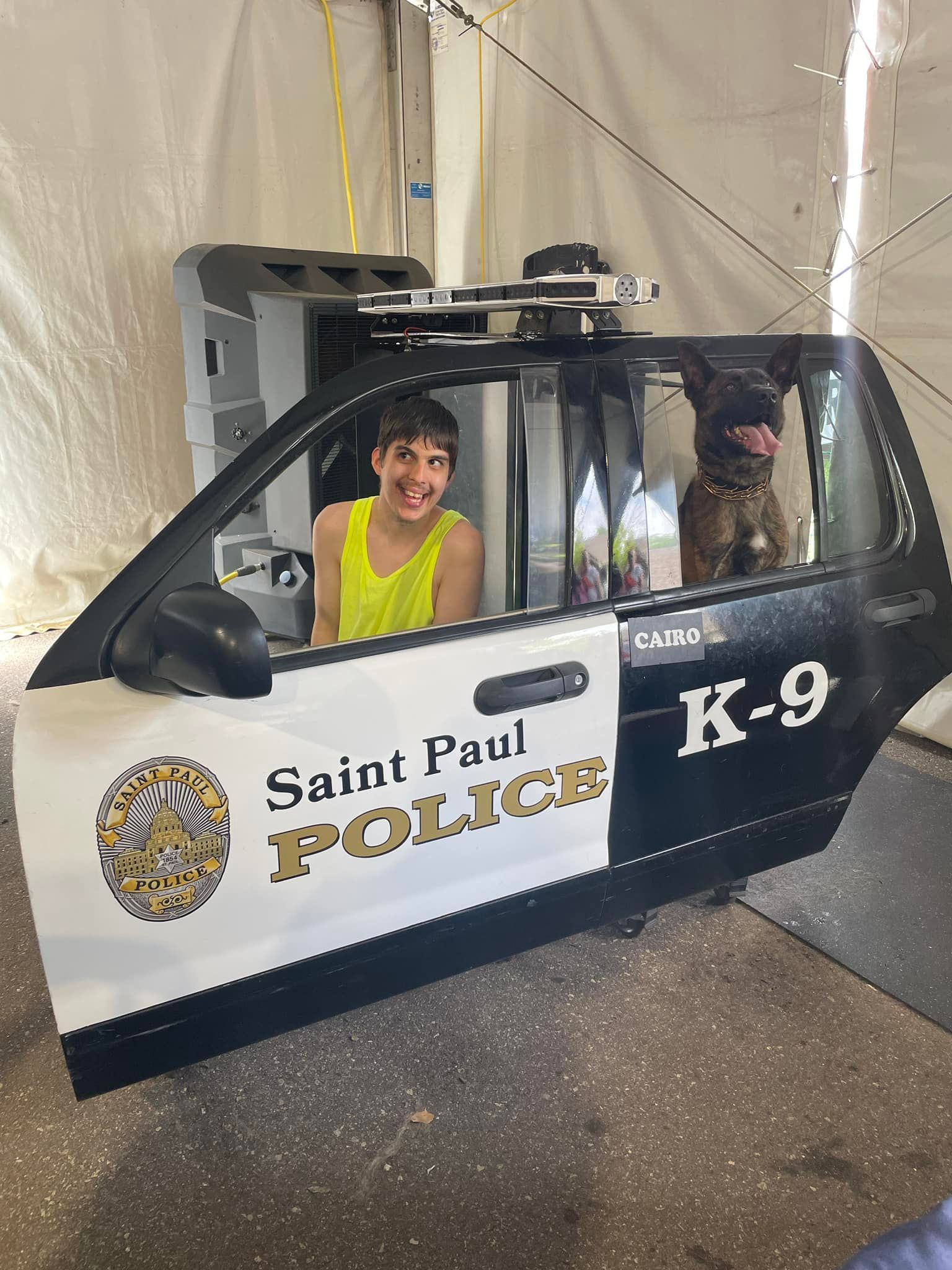 Boy smiles inside a police car door with a K-9 dog. Door says 