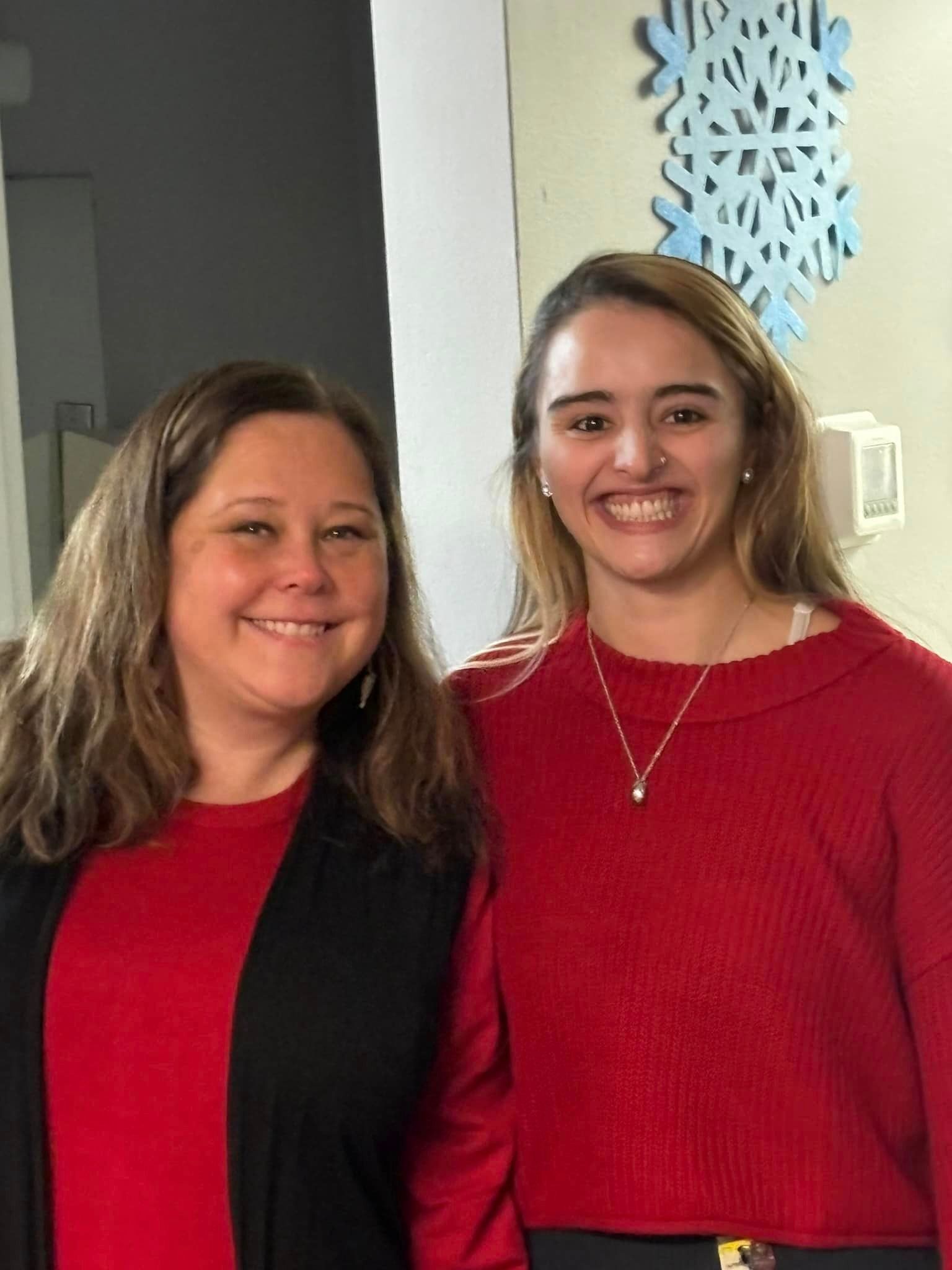 Two women smiling; one in a black cardigan over a red top, the other in a red sweater. Blue snowflake decoration on wall.