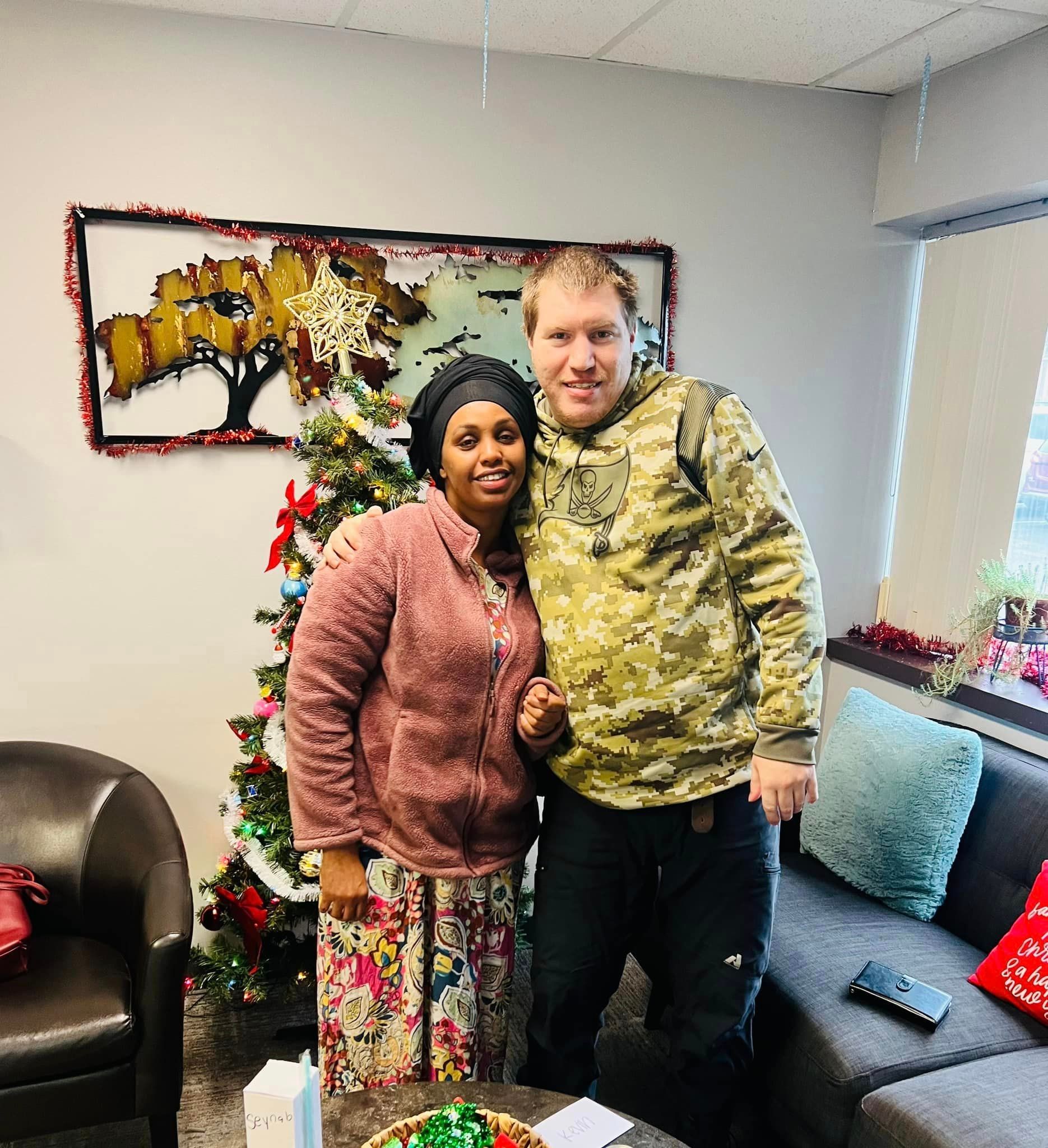 Two people smiling, standing by a decorated Christmas tree in a room with a painting and sofa.