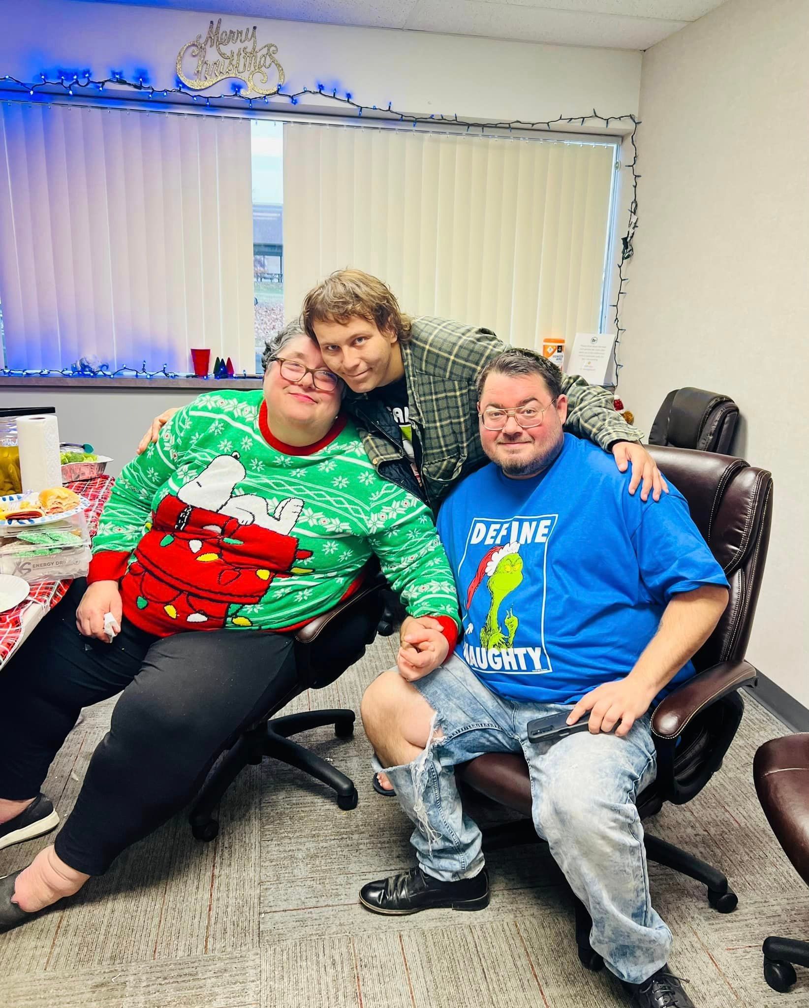 Three people in an office. Two seated, one standing behind. All smiling. One wears a Christmas sweater.