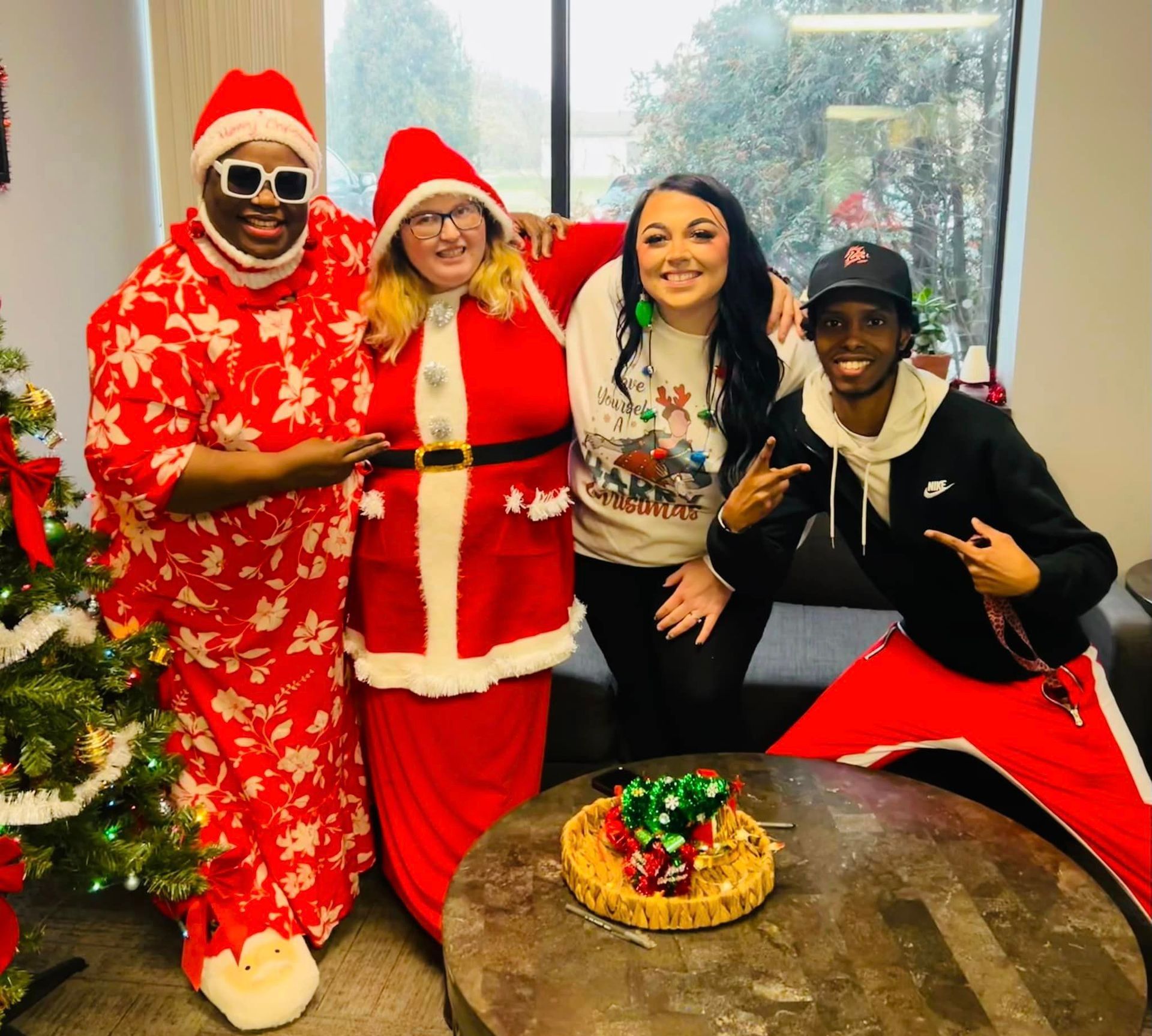 Four people in Christmas attire pose by a decorated tree, two wearing Santa costumes.