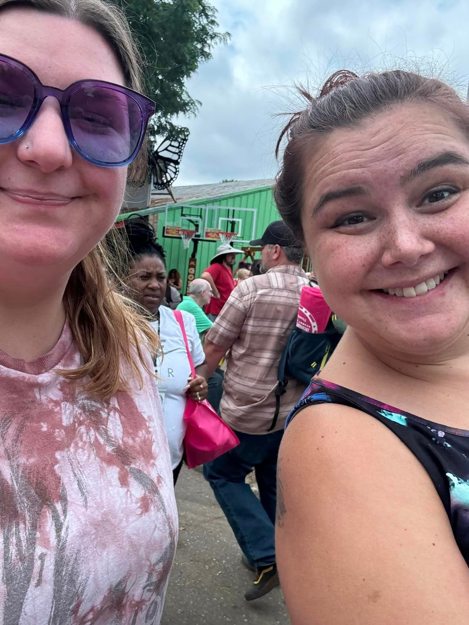 Two smiling women take a selfie in a crowded outdoor setting, people and green structures in the background.
