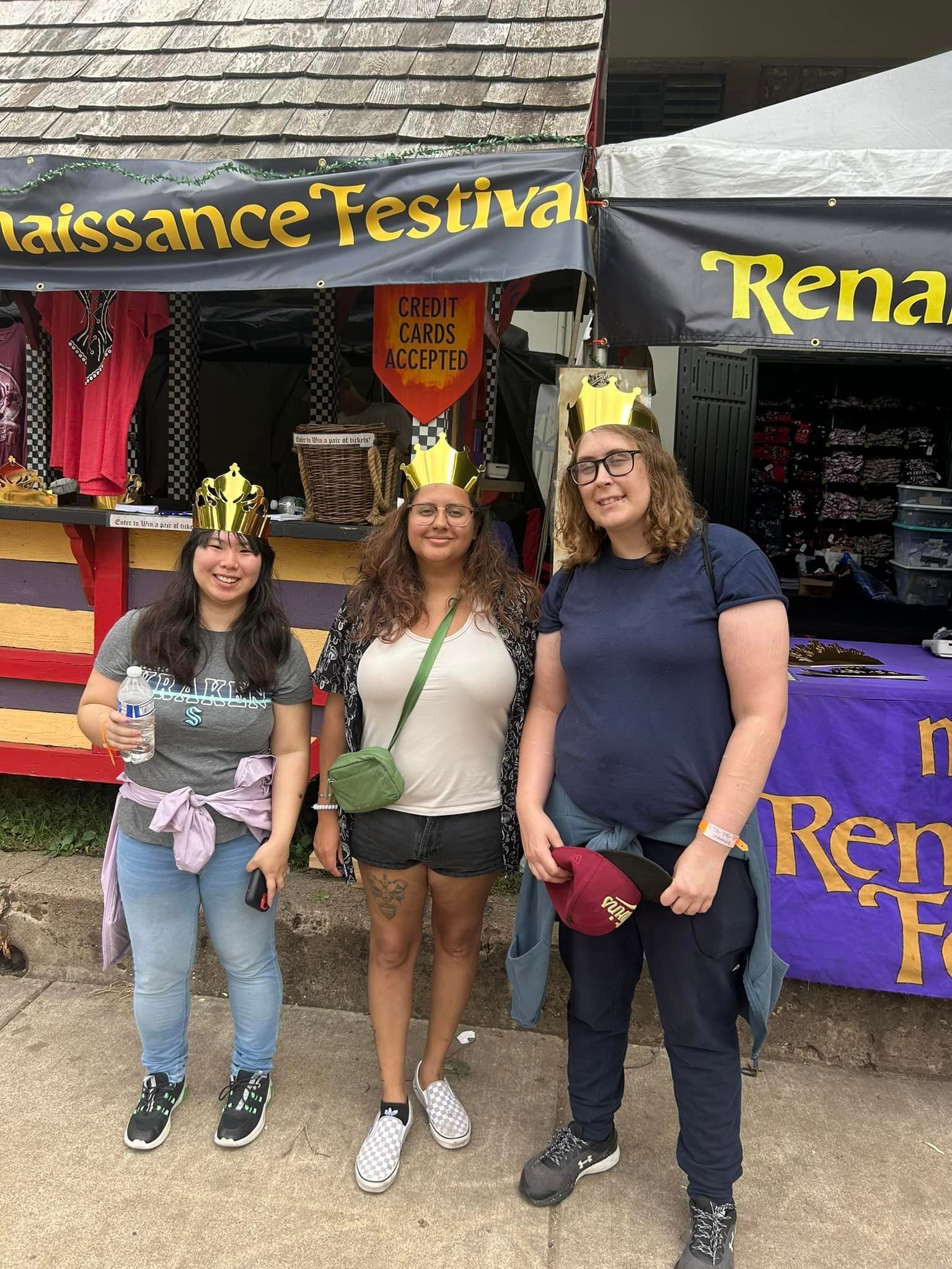 Three people wearing crowns at a Renaissance Festival stand. The background is a vendor's stall.
