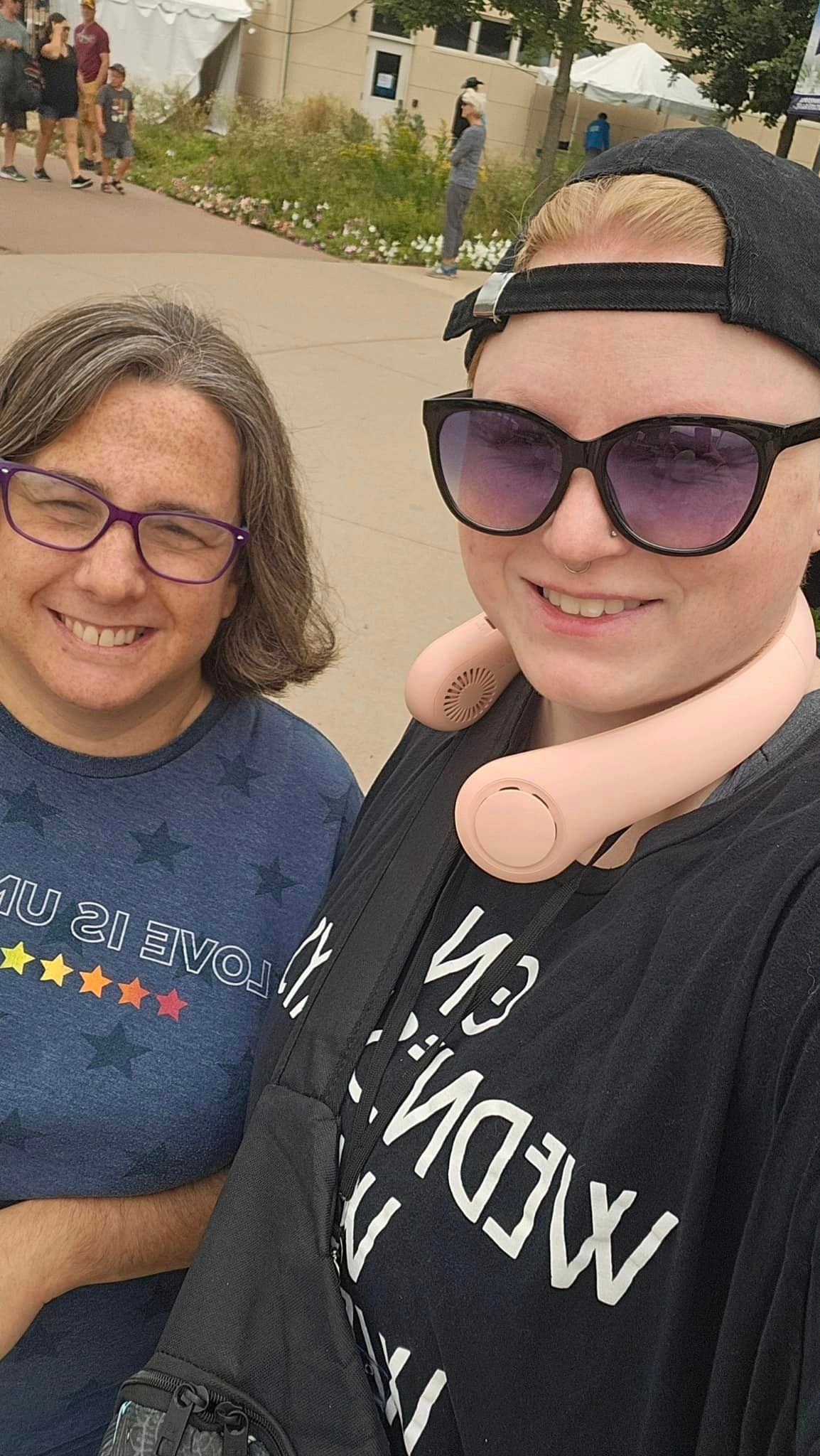 Two people smiling outside. One wears sunglasses, a cap, and a neck fan. The other wears glasses and a starred shirt.