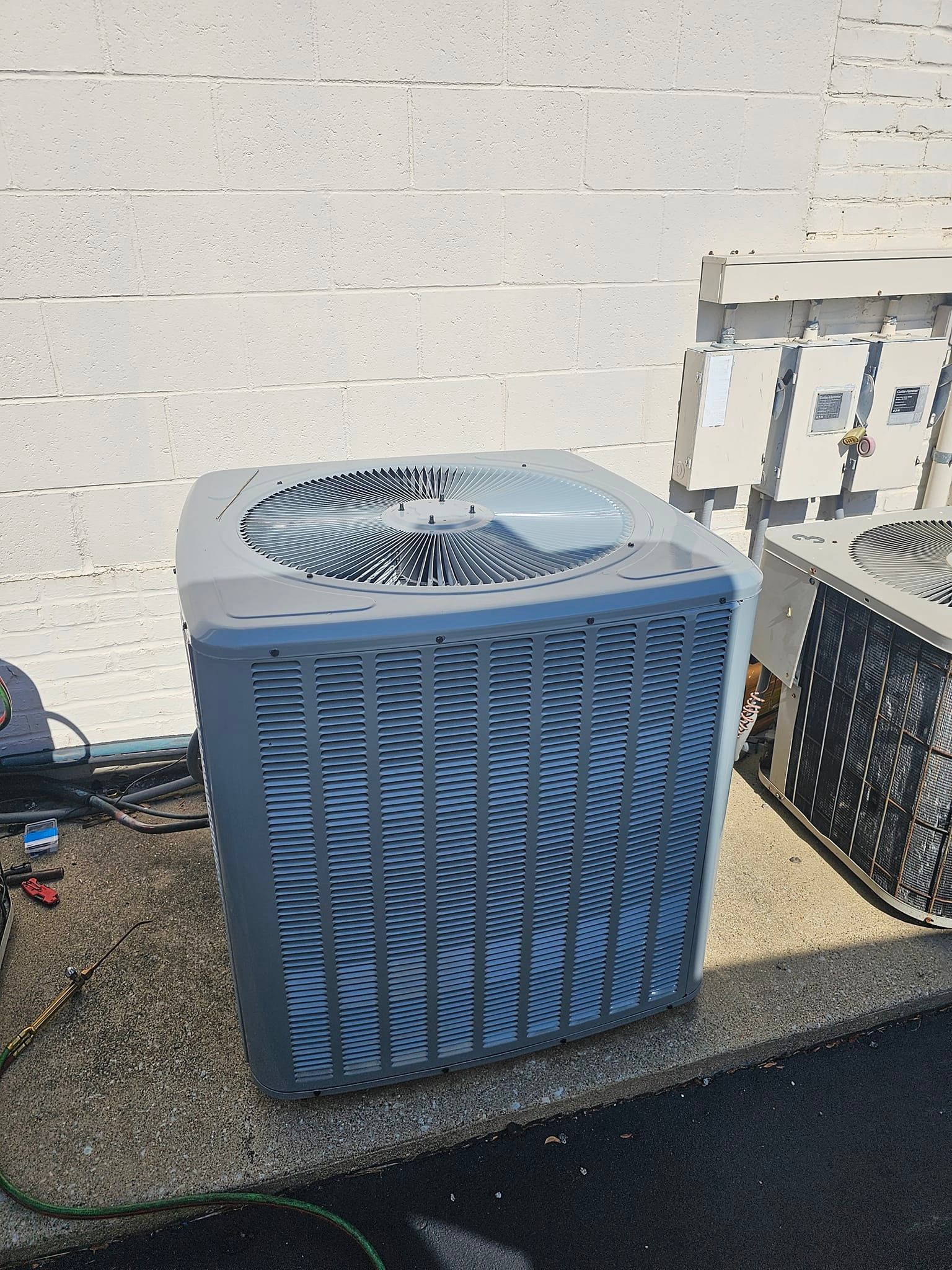 A blue air conditioner is sitting on the sidewalk next to a white brick wall.