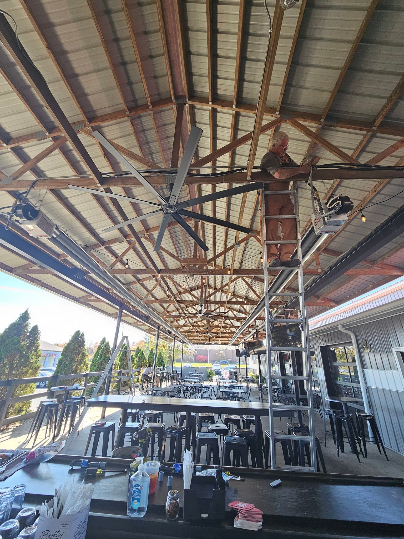 A man is standing on a ladder under a ceiling fan in a restaurant.