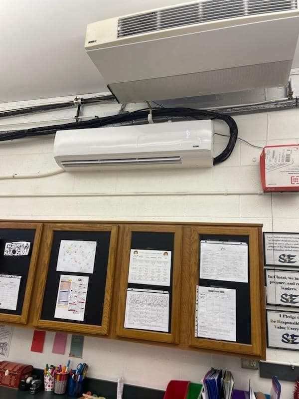 A white air conditioner is hanging from the ceiling above a bulletin board