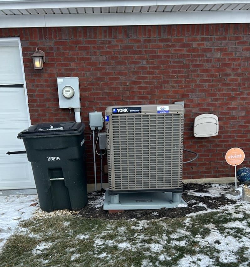 A black trash can is sitting next to a large air conditioner on the side of a brick house.