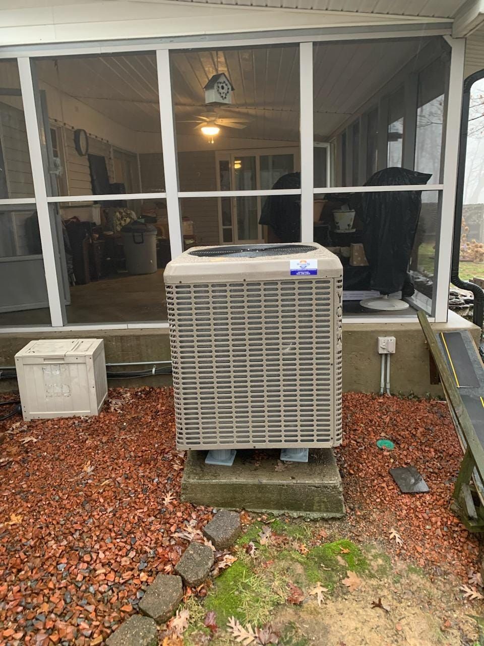 A large air conditioner is sitting on top of a pile of gravel in front of a screened in porch.