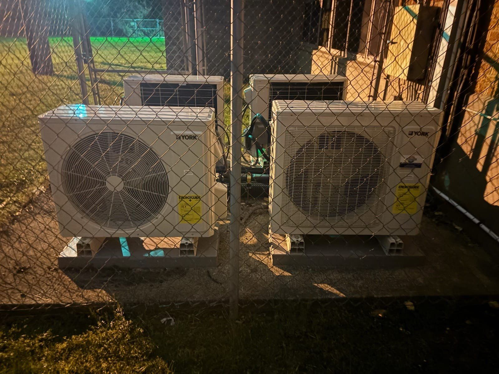 Two air conditioners are sitting next to each other in front of a chain link fence.