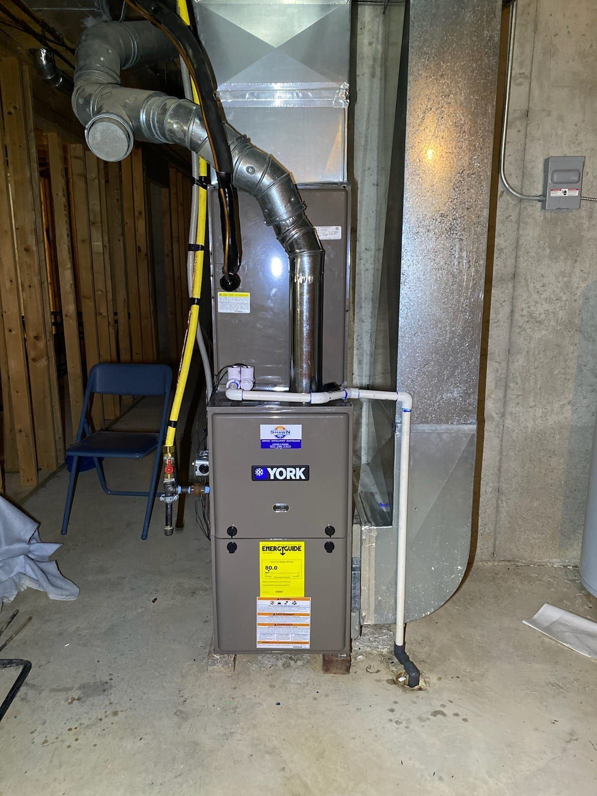 A york furnace is sitting in a basement next to a chair.