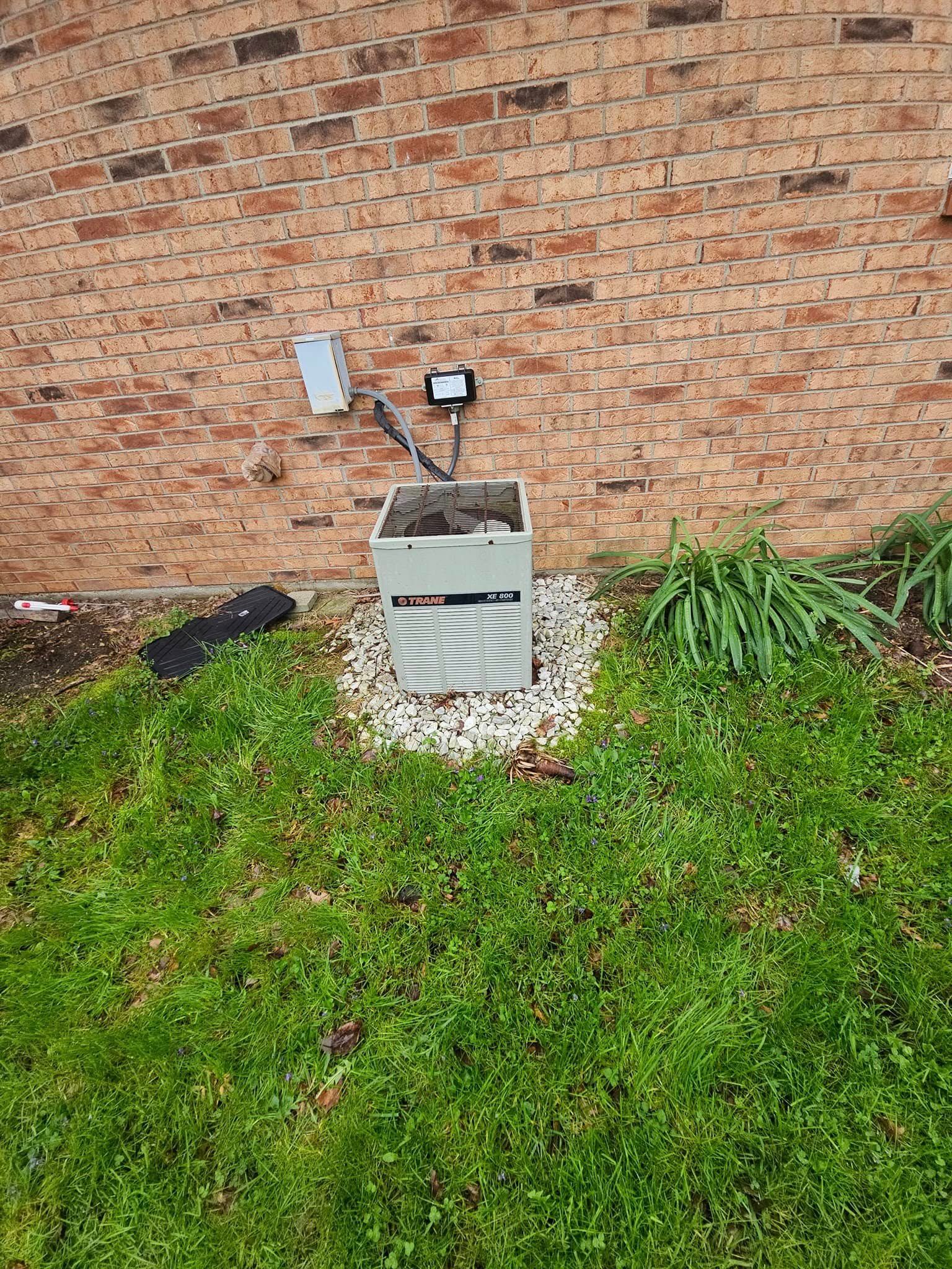 A generator is sitting in the grass in front of a brick wall.
