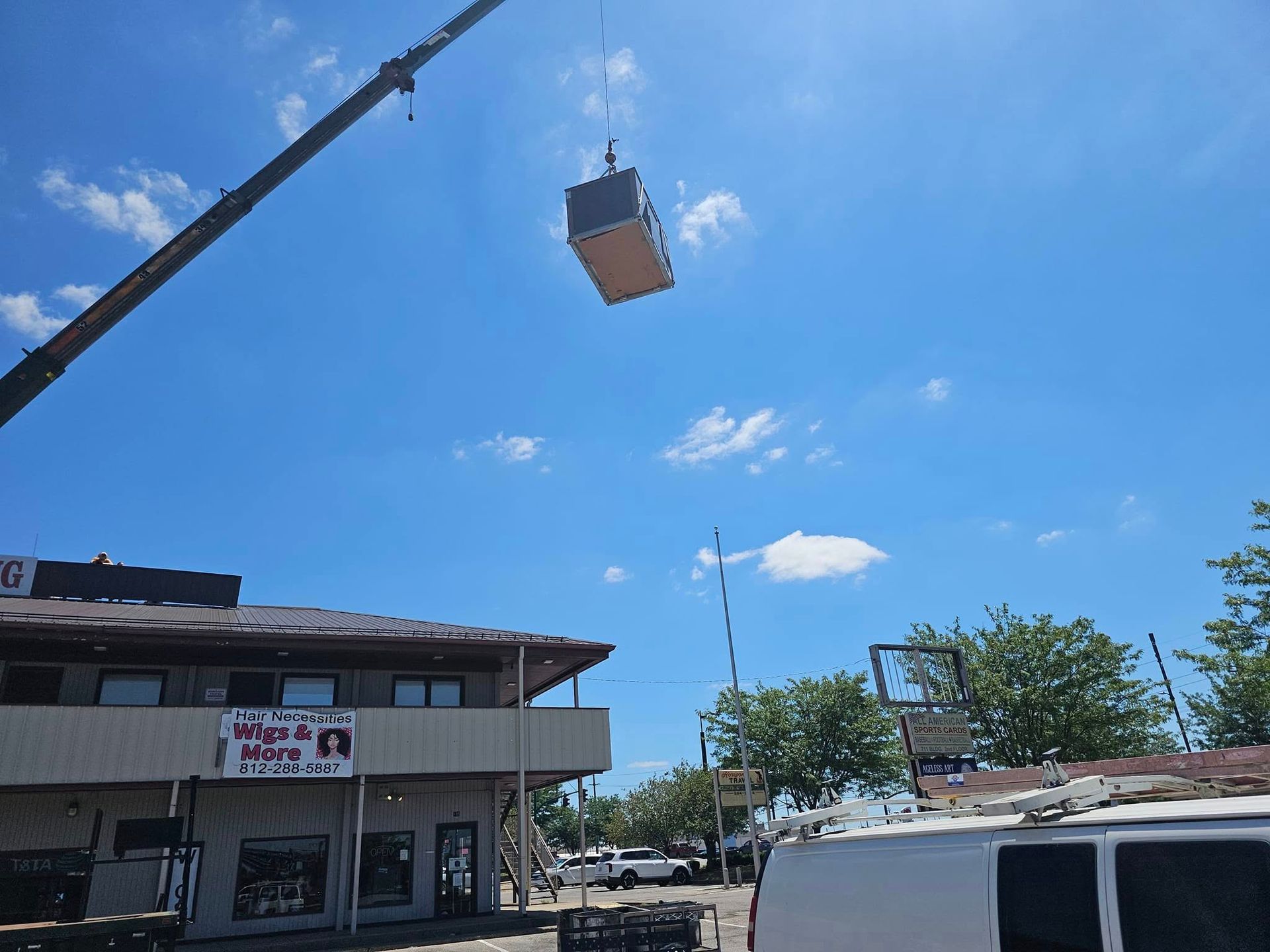 A large box is being lifted by a crane in front of a building with a sign that says wigs & more