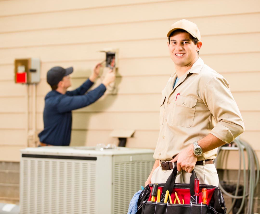 A man with a tool bag is standing in front of an air conditioner while another man works on it.