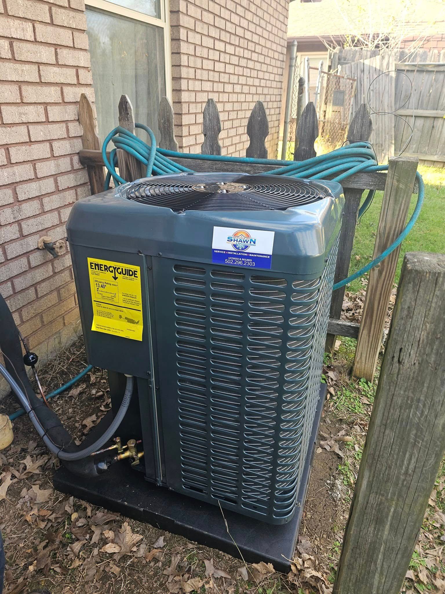 An air conditioner is sitting on the side of a brick building next to a fence.
