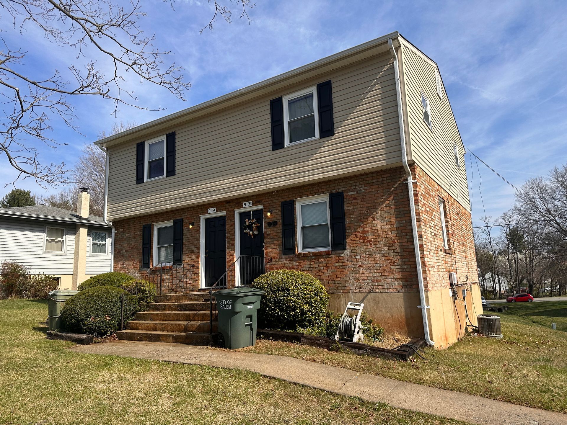 Two-story brick and tan-sided duplex with black shutters, green bushes, and a green trash can in front. Roanoke