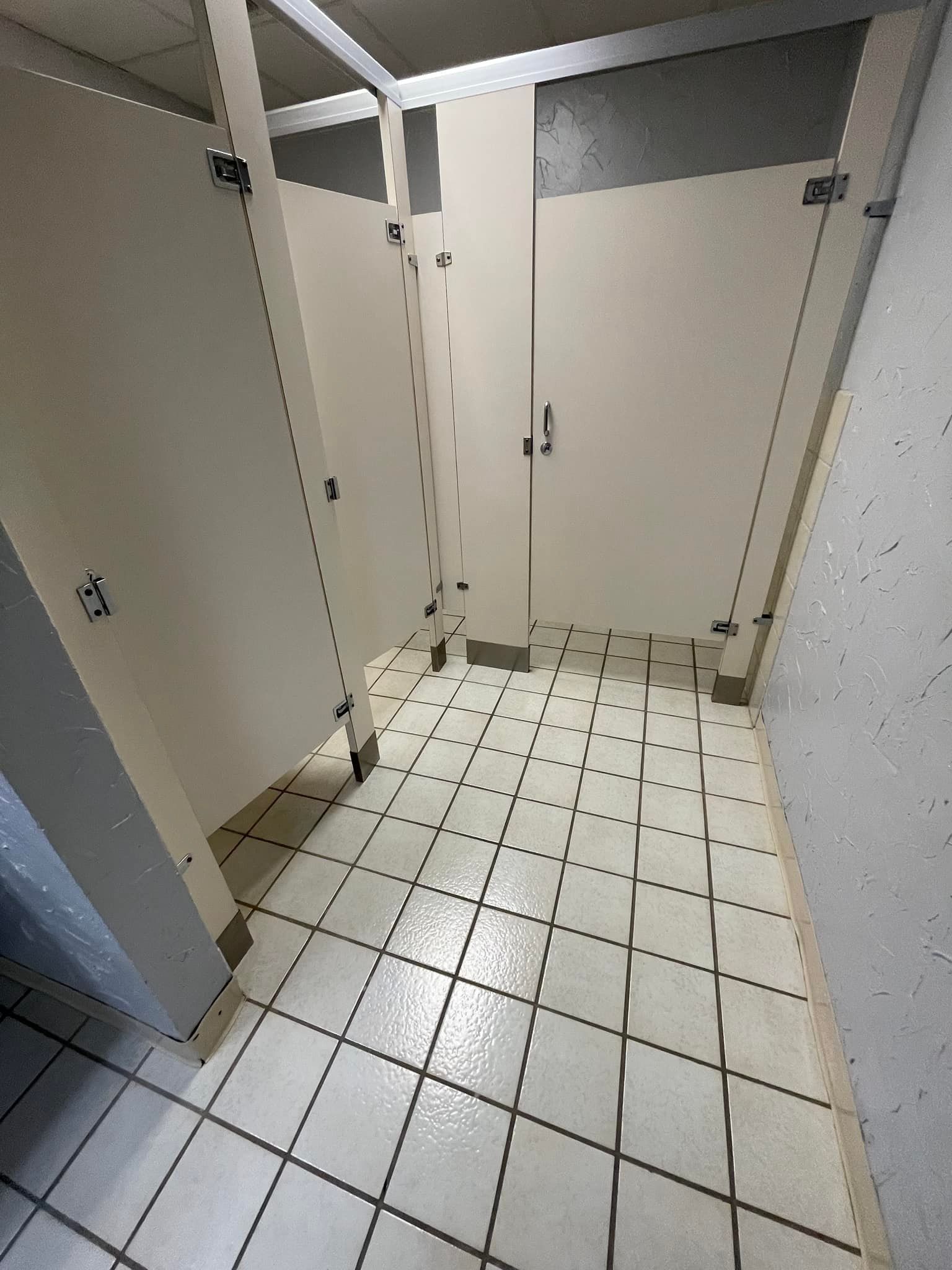 Three public restroom stalls with off-white doors, white tile floor, and metal fixtures.