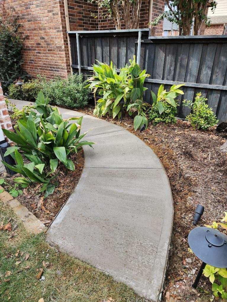 Concrete walkway curves through a garden bed with green plants and brown mulch. Brick building & a wooden fence are present.