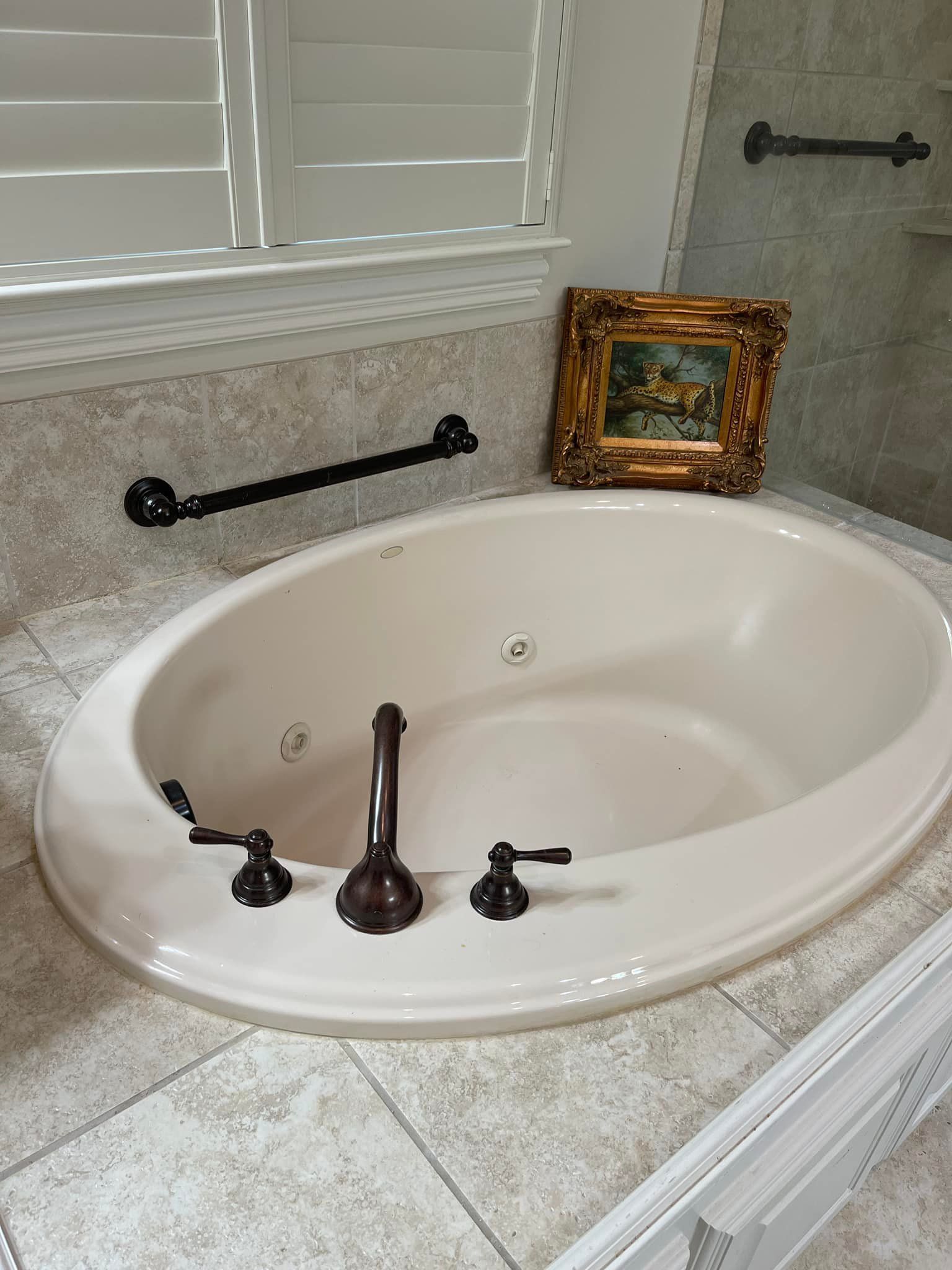 Oval bathtub with oil rubbed bronze fixtures, near a window with shutters, and a small framed painting.