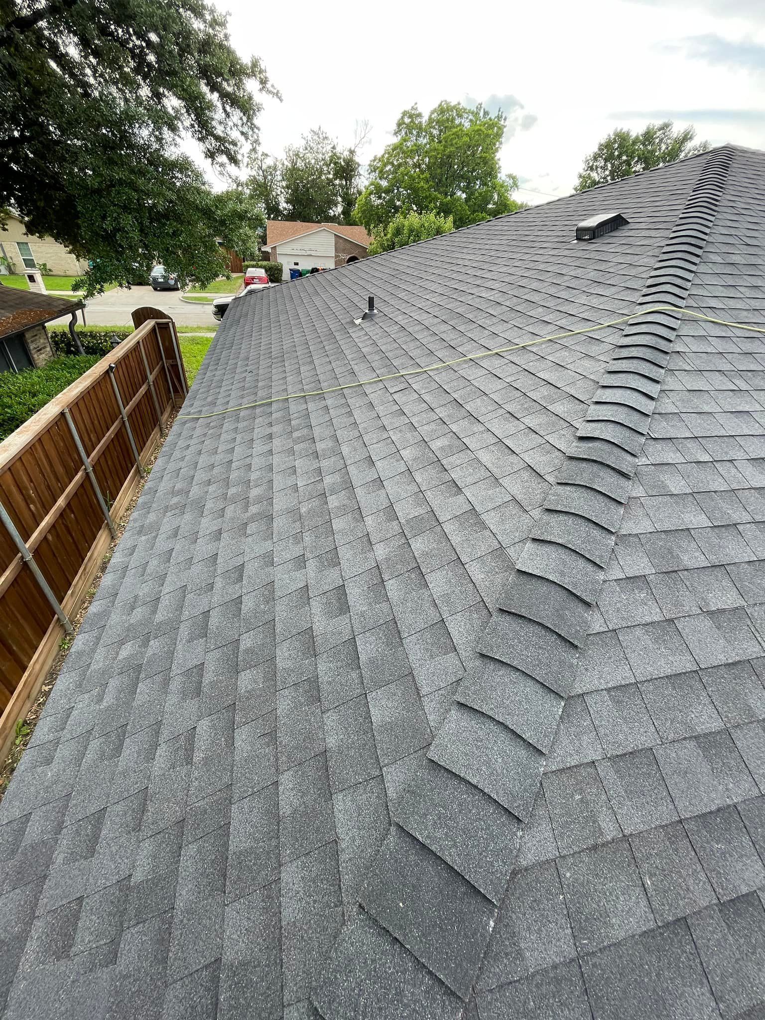 Roof covered with gray asphalt shingles, viewed from above beside a wooden fence and trees