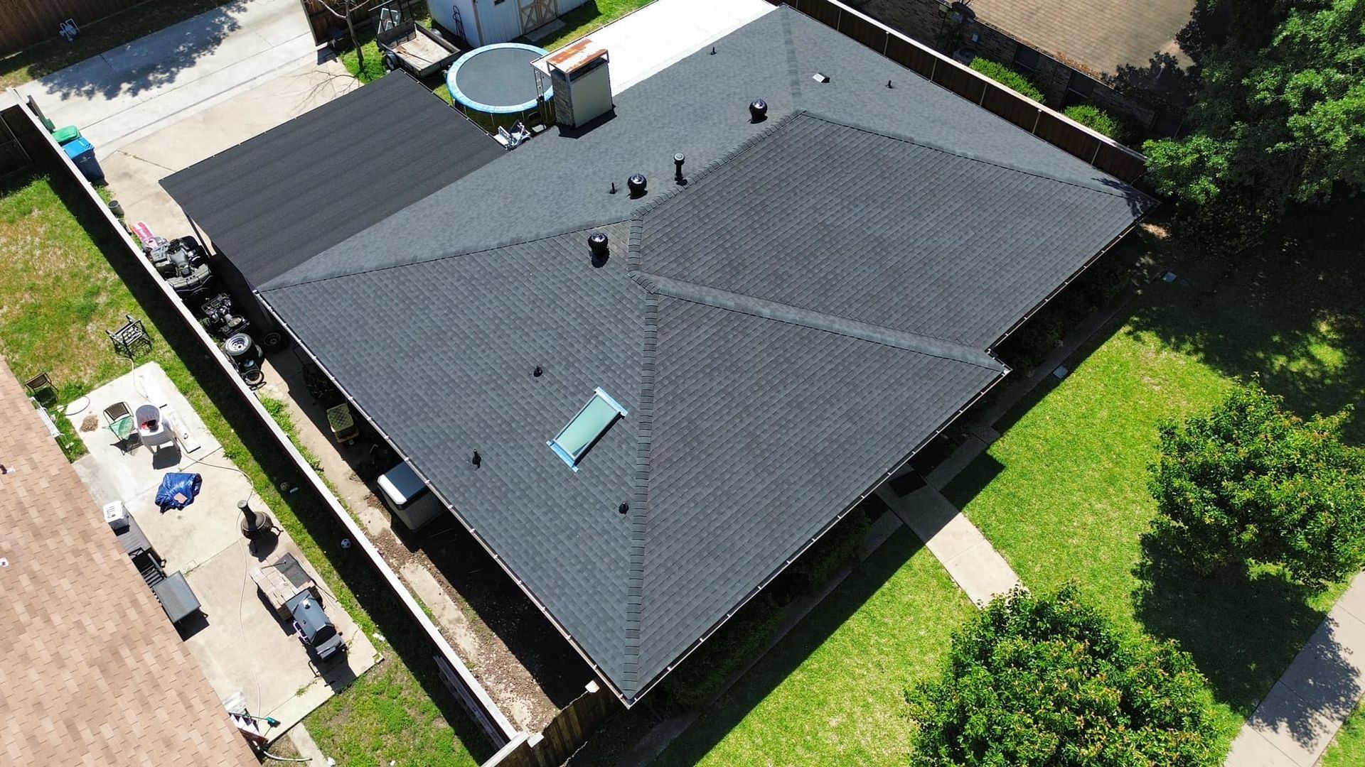 Aerial view of a dark shingled roof with a chimney, skylight, and surrounding yard with trees.