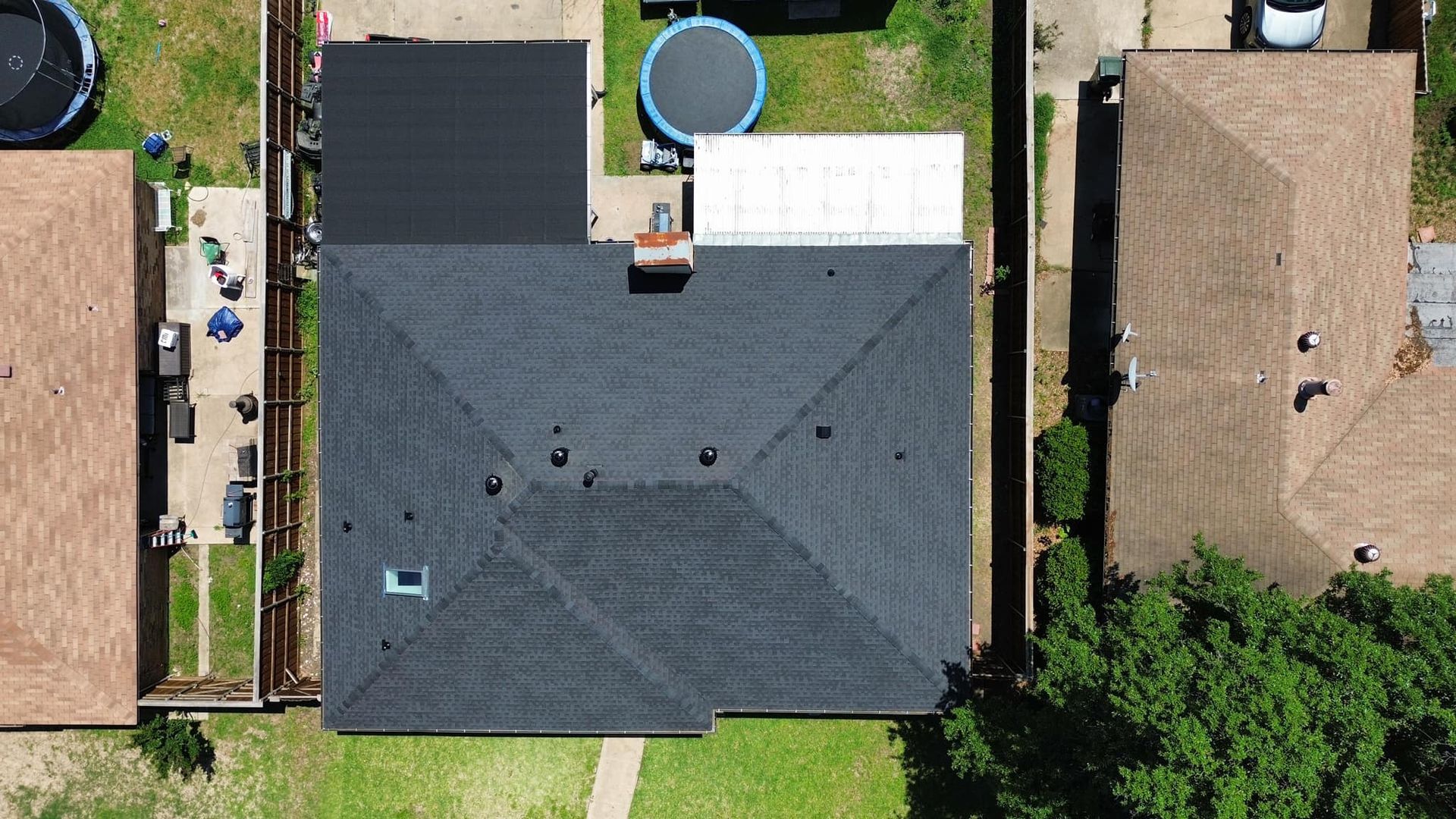 Aerial view of a house with a black roof, a small trampoline, and surrounding houses with grass.