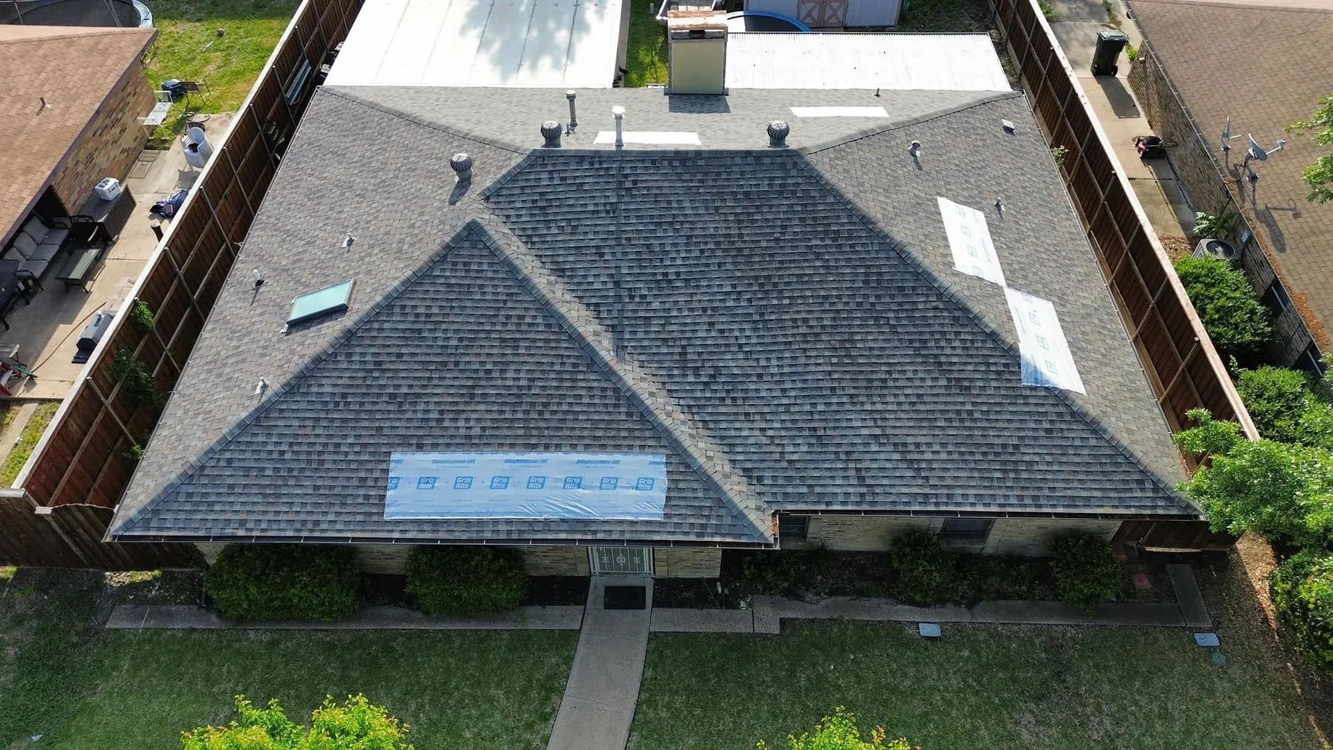 Aerial view of a gray shingled roof on a home, with a sign visible, surrounded by a brown fence.