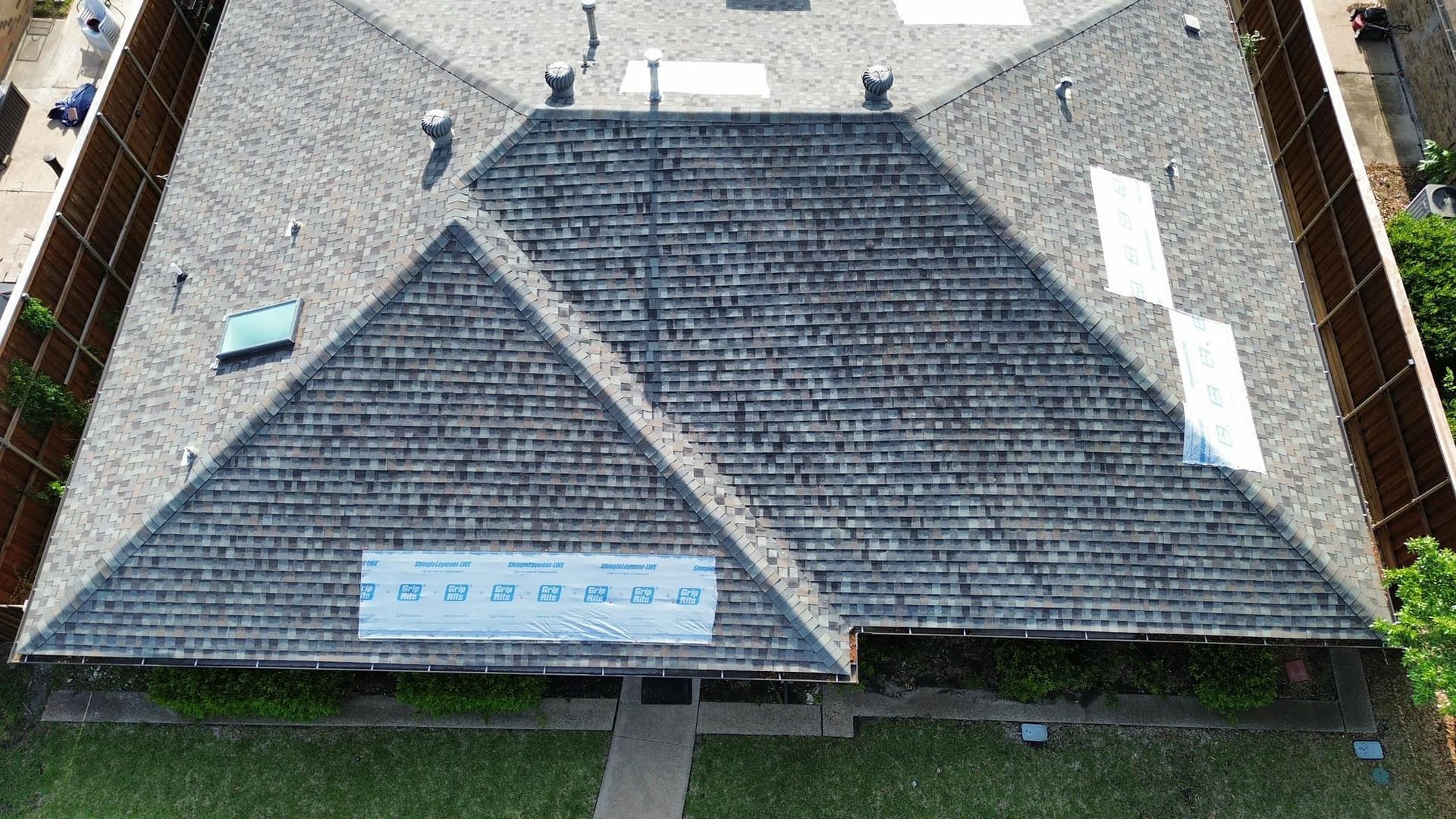 Overhead view of a building with a gray, multi-sectioned roof, surrounded by a brown fence and green lawn.