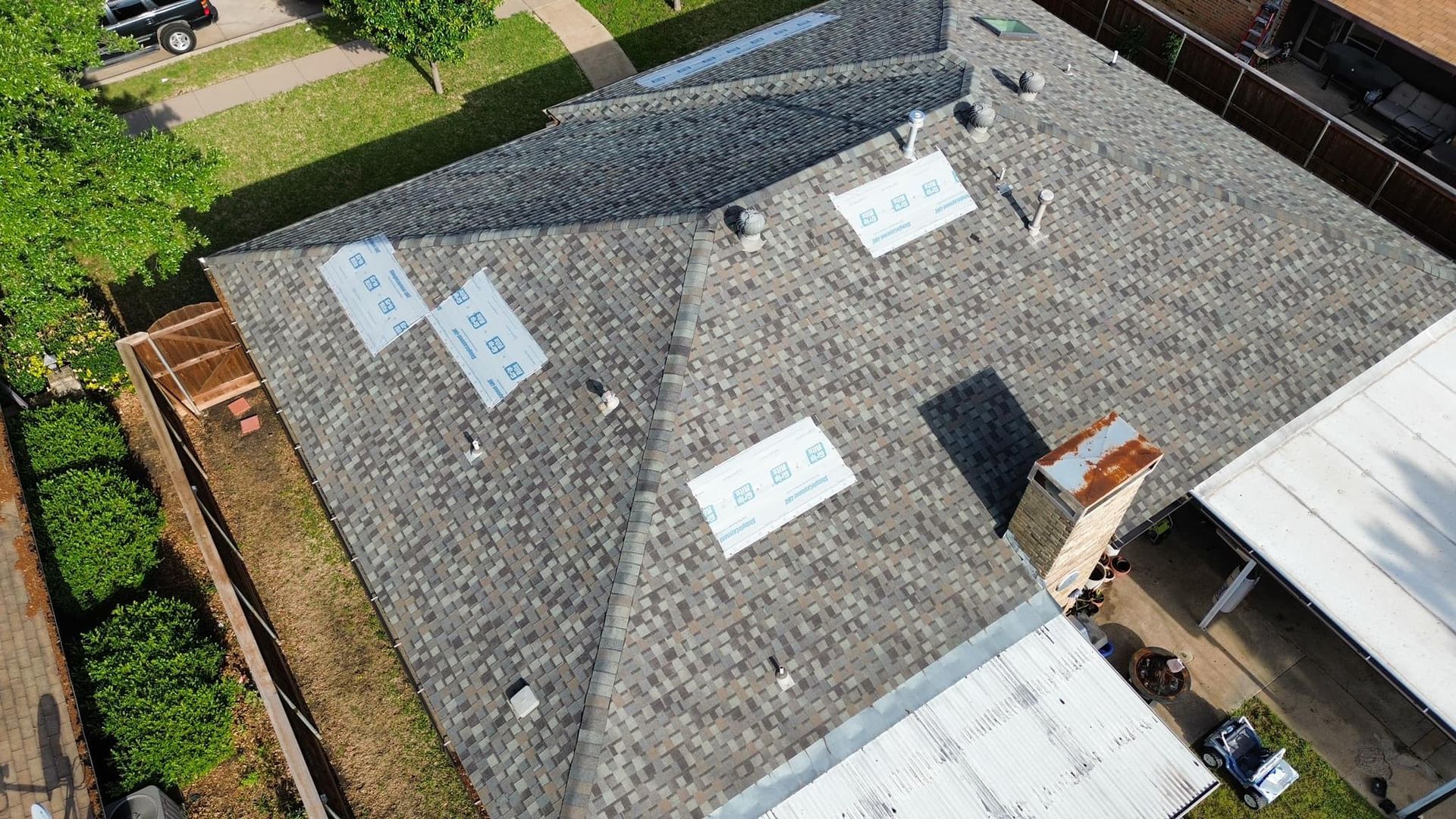 Aerial view of a gray shingle roof with several rectangular vents and a chimney, viewed from above.