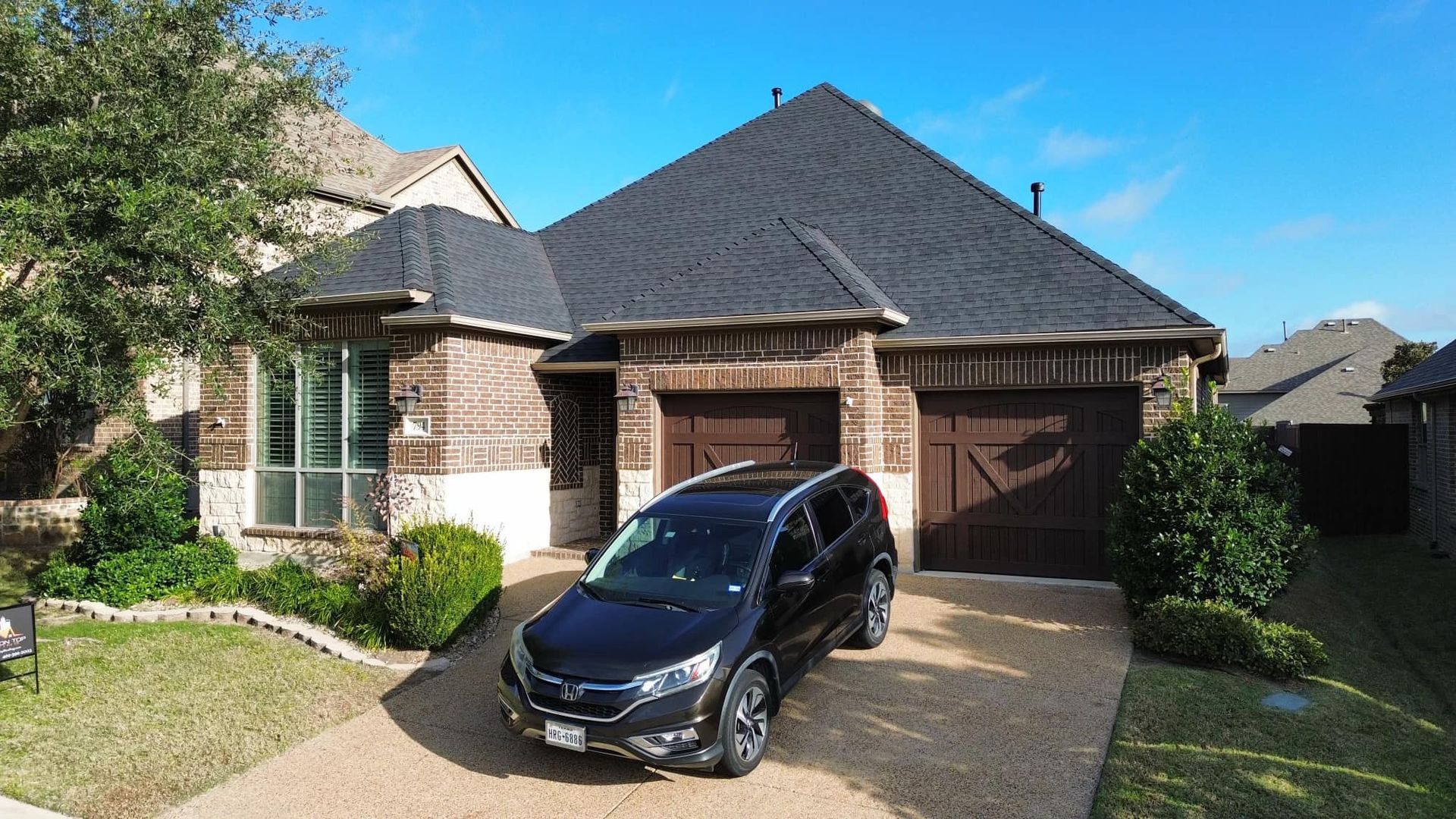 Black car parked in front of a brick house with a dark roof and two car garage on a sunny day.