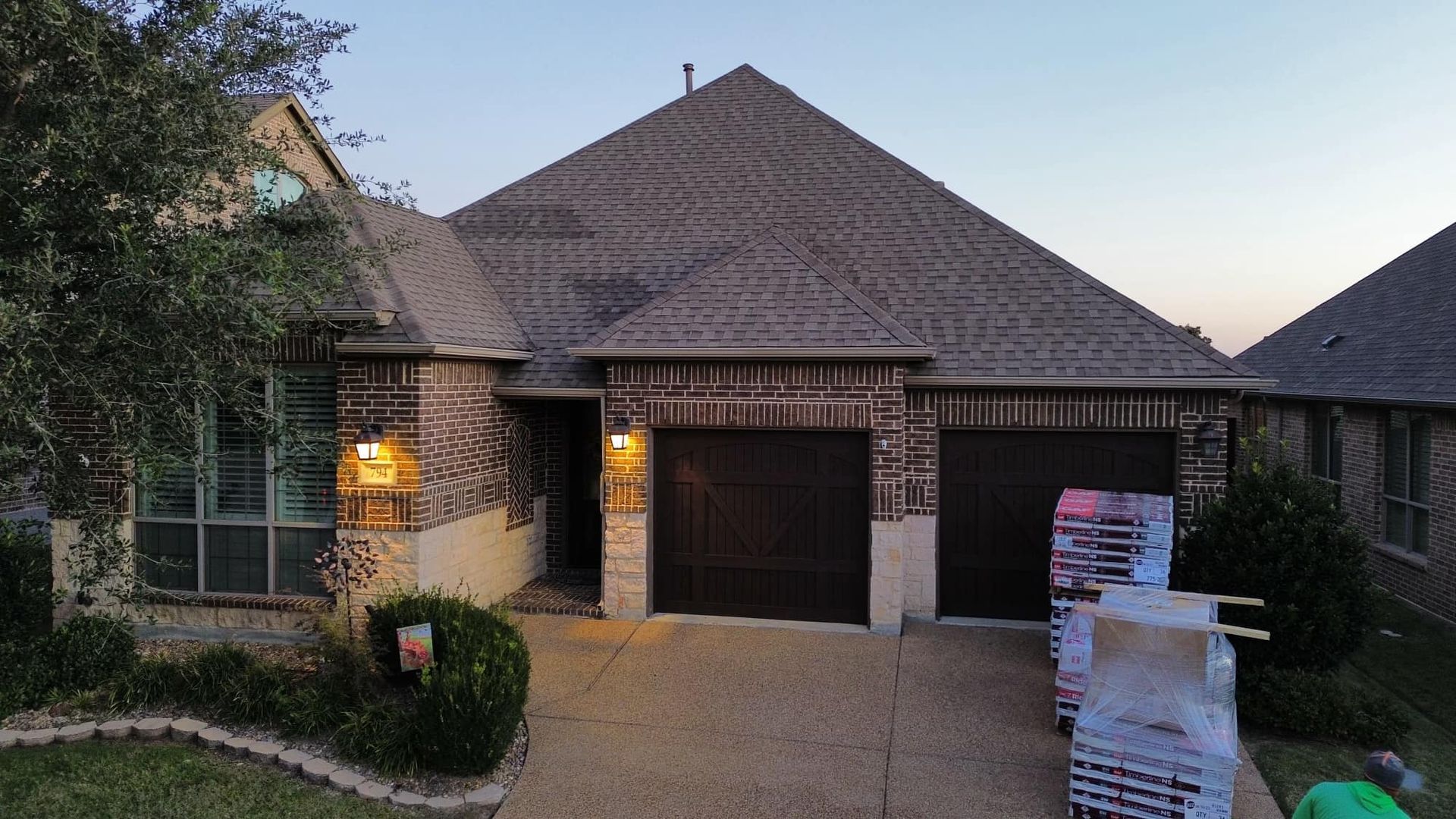 Brick house with brown roof and garage doors, lit by outdoor lights, dusk setting.