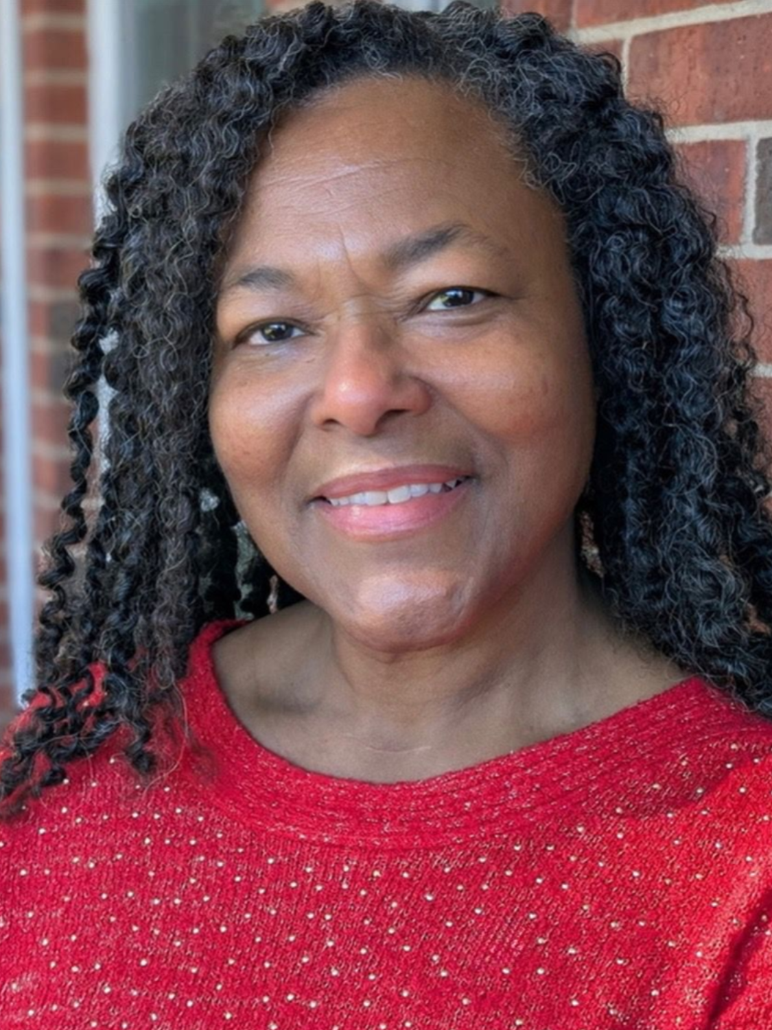Woman with dark curly hair smiles, wearing a red sweater, standing in front of a brick wall.